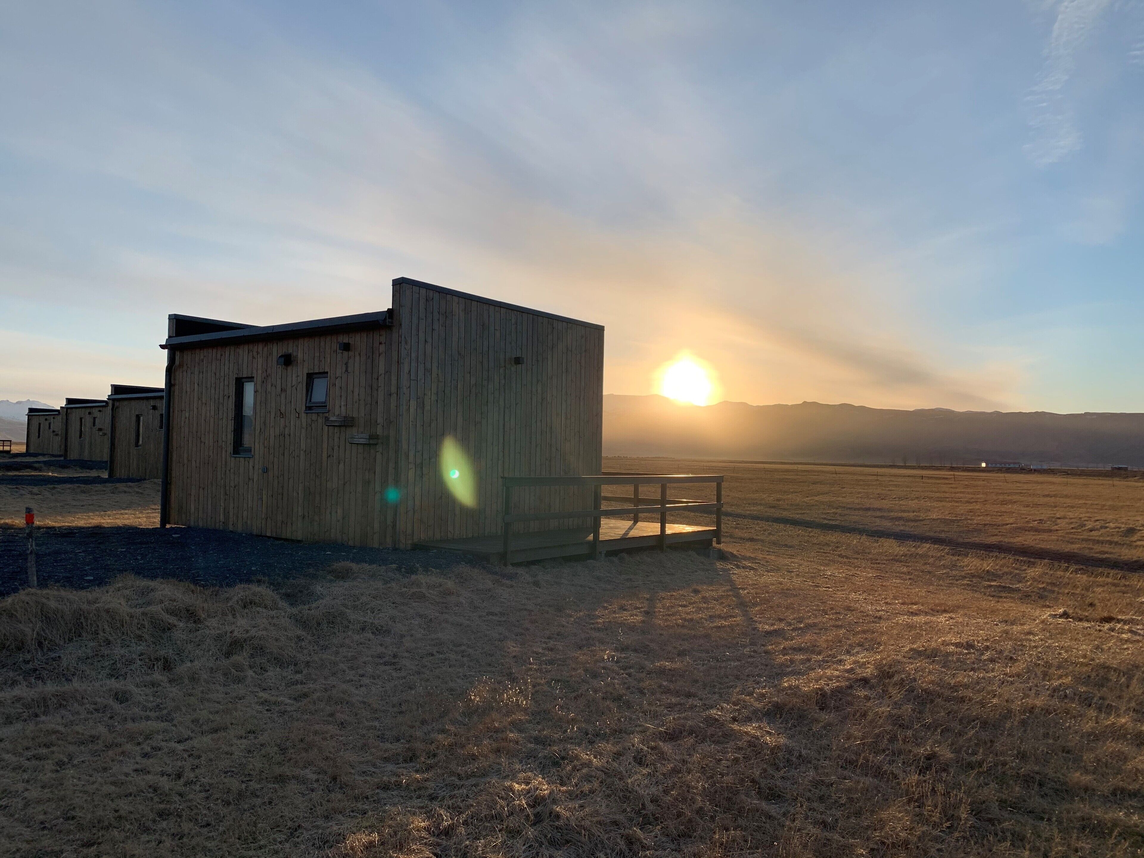 A wooden cabin with a bench, set against a backdrop of a sunset over a flat landscape.