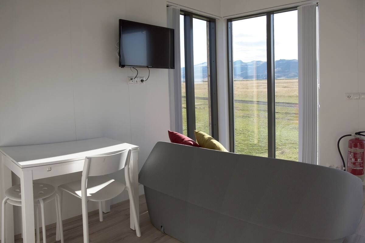A room with a white desk, two white chairs, a grey sofa, a mounted TV, and a large window with a view of a grassy field and mountains.