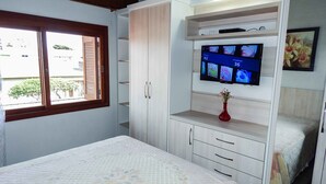 Un dormitorio con cama, televisor montado en un mueble, ventana y un florero sobre la cómoda.