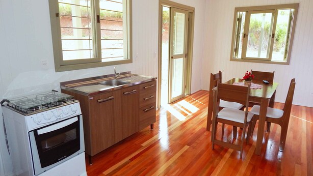 Cocina con estufa a gas, pileta y armarios de madera. Hay una mesa de comedor con sillas y una ventana con persianas.