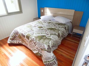 A bedroom with a wooden floor, a bed with a patterned bedspread, a nightstand, and a window with blinds.