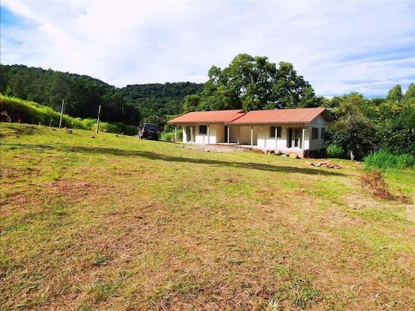 A single-story house with a red roof, surrounded by a grassy area and a forested hill.