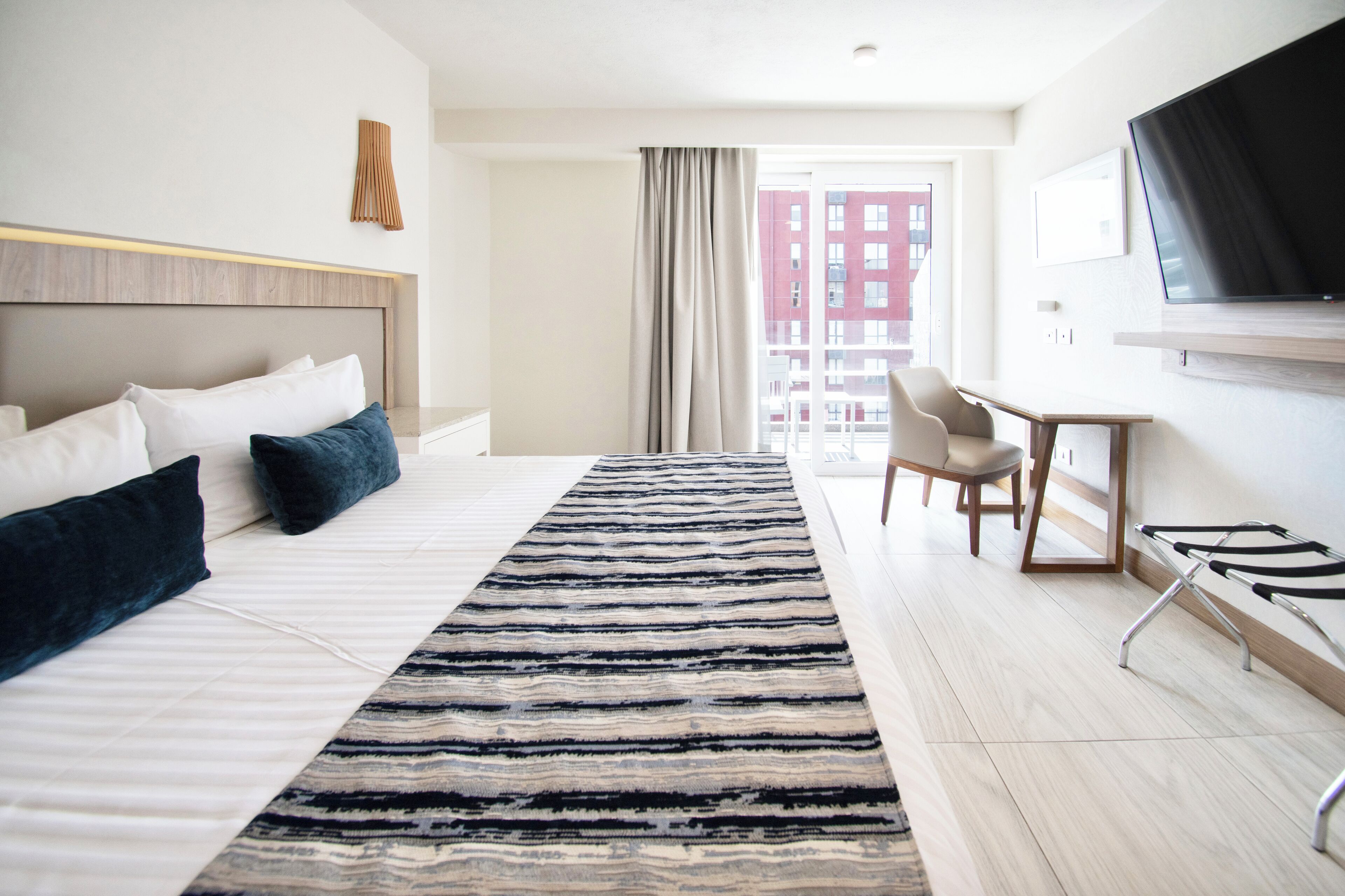 A hotel room with a bed, a TV, a desk, and a view of buildings outside.