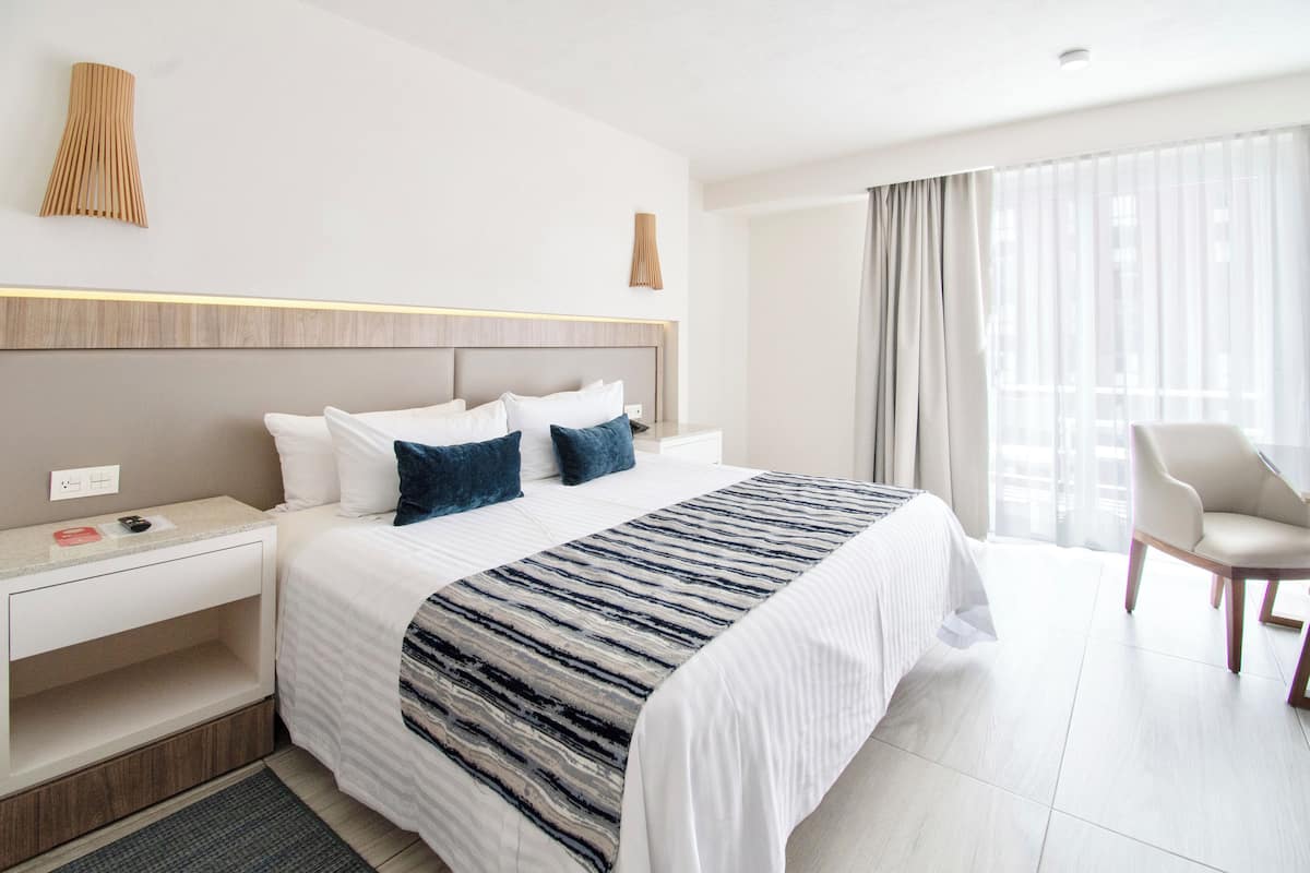 Una habitación de hotel moderna con una cama grande, una mesita de noche, una silla y un ventanal con cortinas.