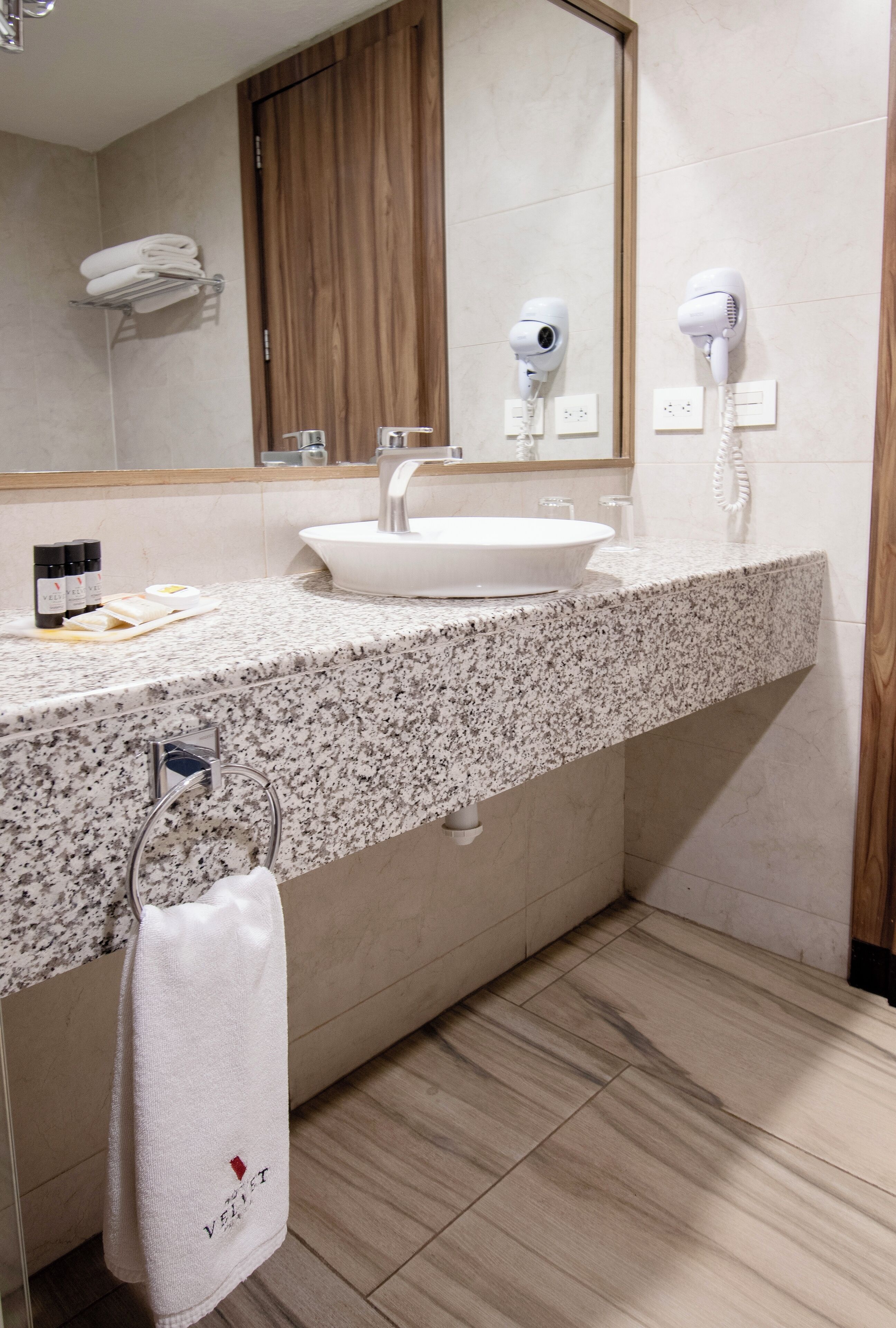 Un baño con encimera de granito, un lavabo blanco, un espejo y dos secadores de pelo fijados en la pared.