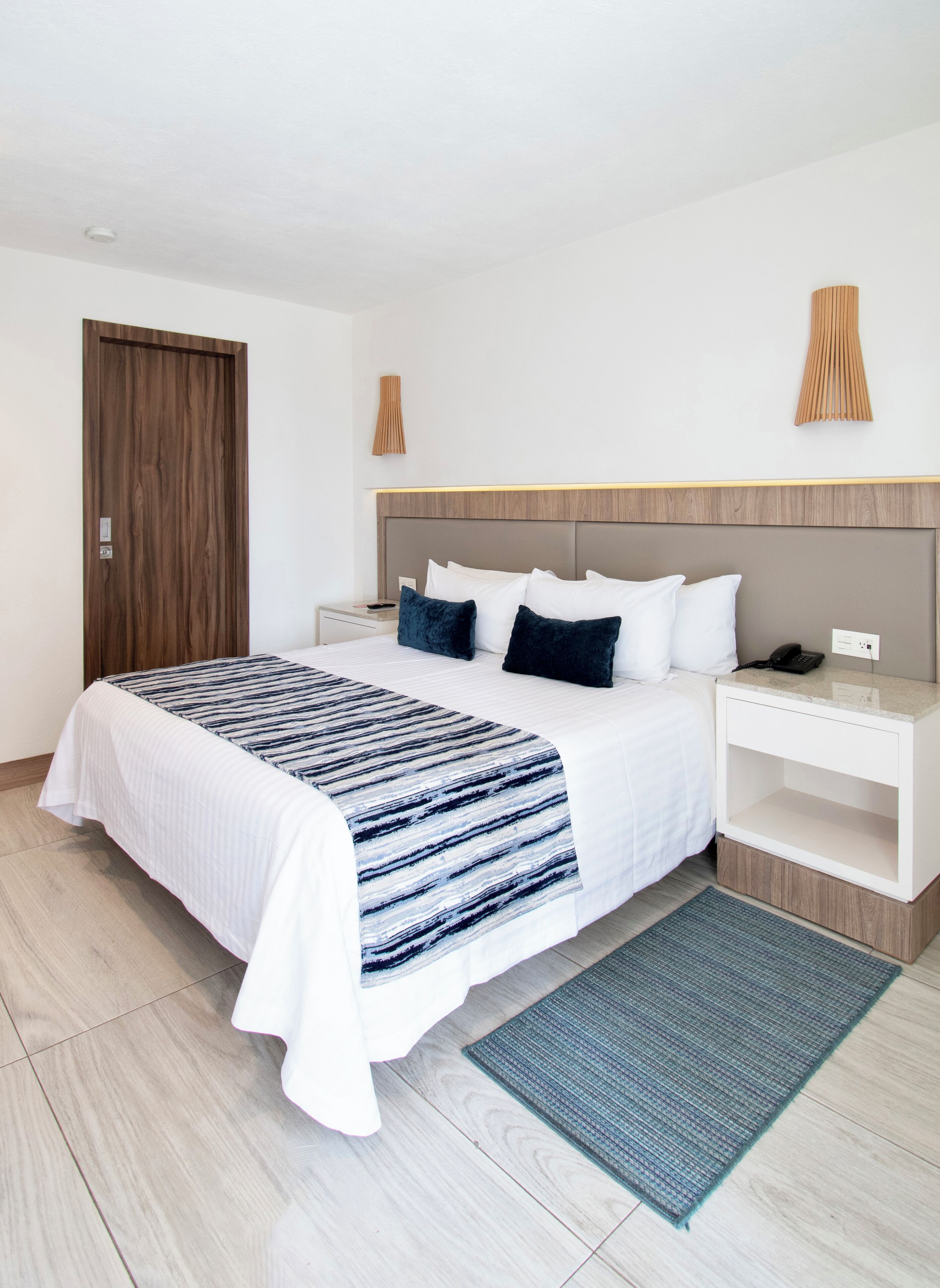 A modern hotel room with a large bed, wooden headboard, white nightstands, and a wooden door.