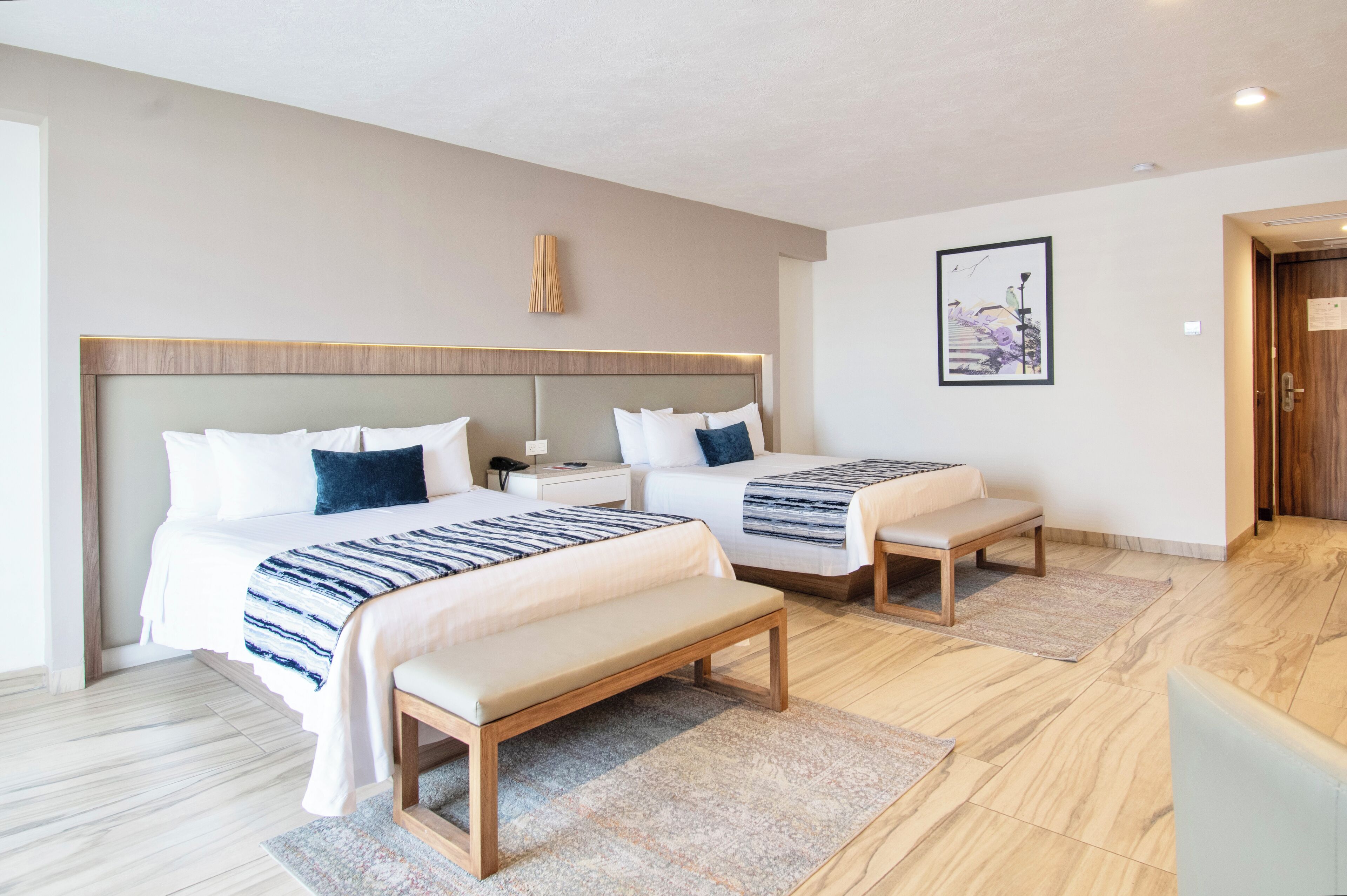 Camera d'albergo con due letti, una panca, un comodino, una lampada e un quadro appeso alla parete.