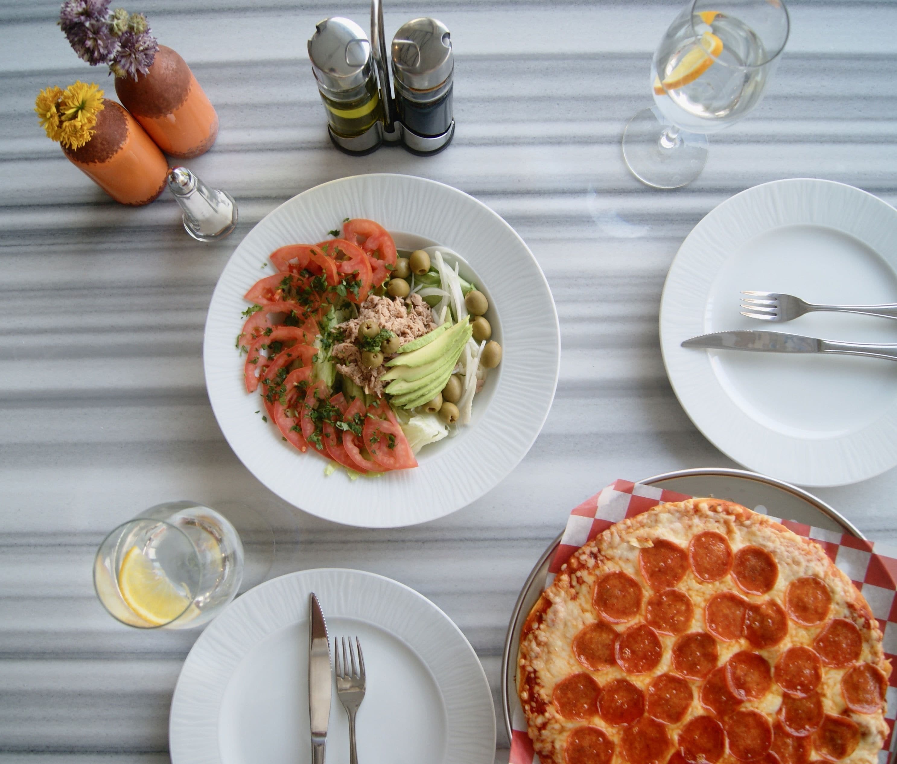 A table with plates of food, a pepper shaker, salt and pepper shakers, a glass of water, and a pizza.