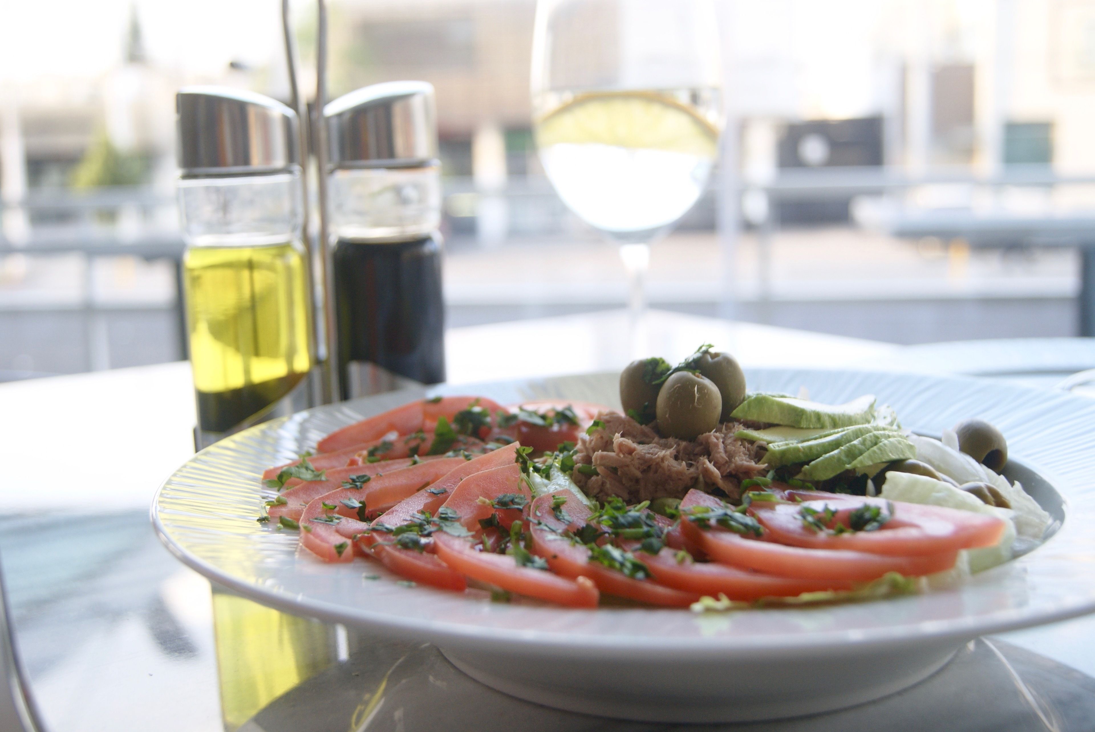 A plate of food with tomatoes, olives, and greens, accompanied by two bottles of olive oil and a glass of white wine on a table.
