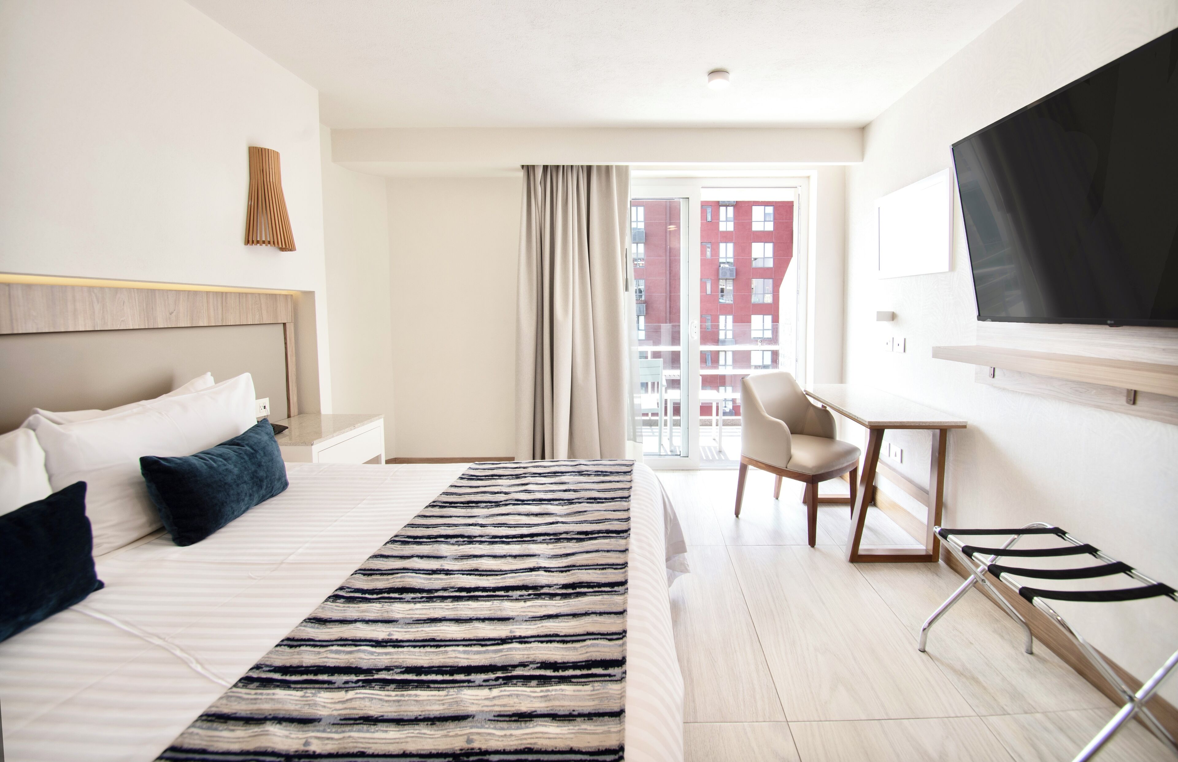 A hotel room with a bed, a desk, a chair, a TV, and a view of buildings through the window.