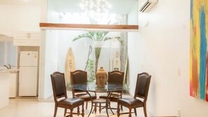 Un coin repas avec une table en verre entourée de chaises en bois, un lustre au-dessus, et une vue sur une cuisine et une plante, le tout visible à travers la vitre.
