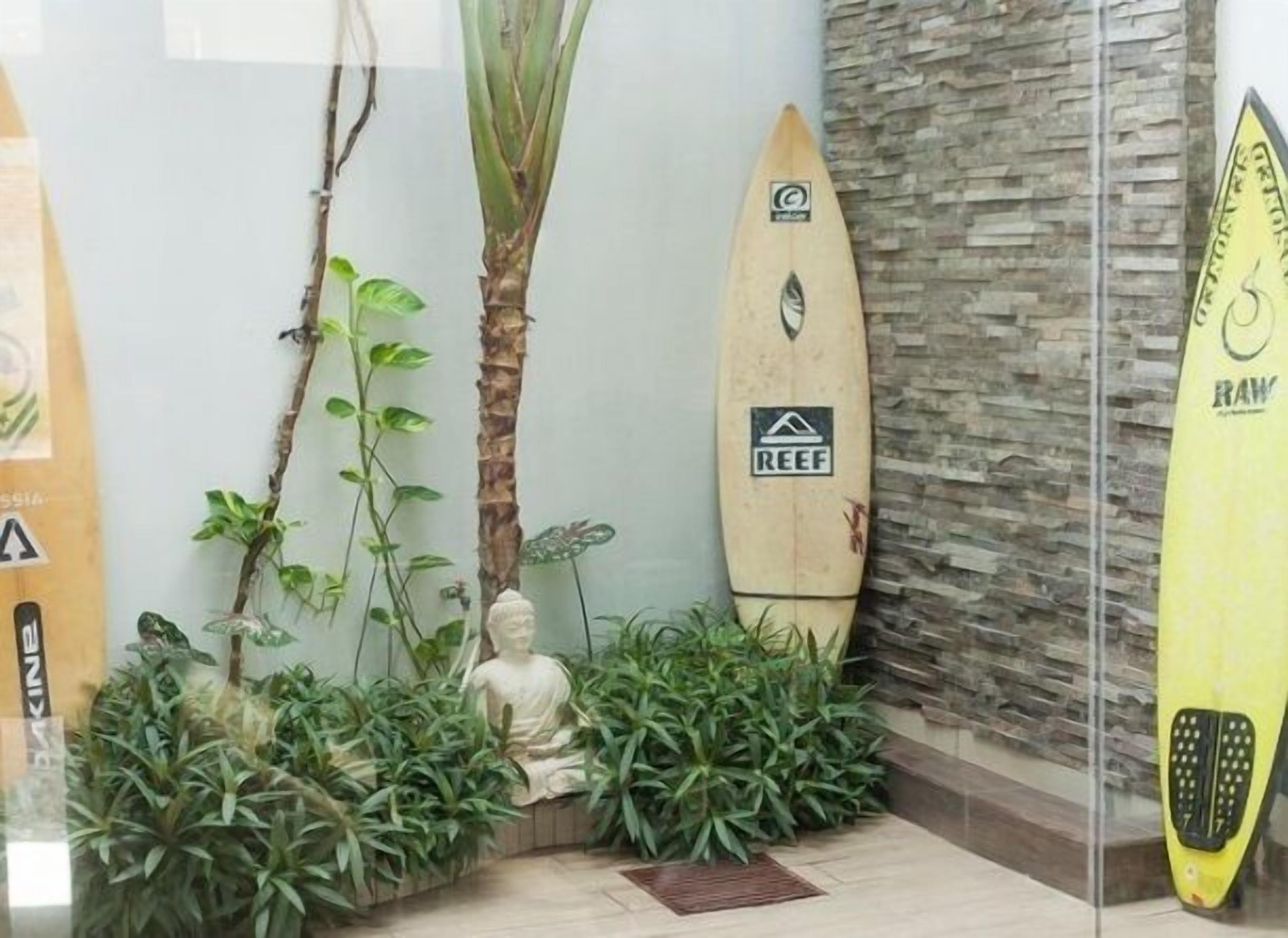 Une pièce avec une cloison en verre, une planche de surf, une statue et diverses plantes.