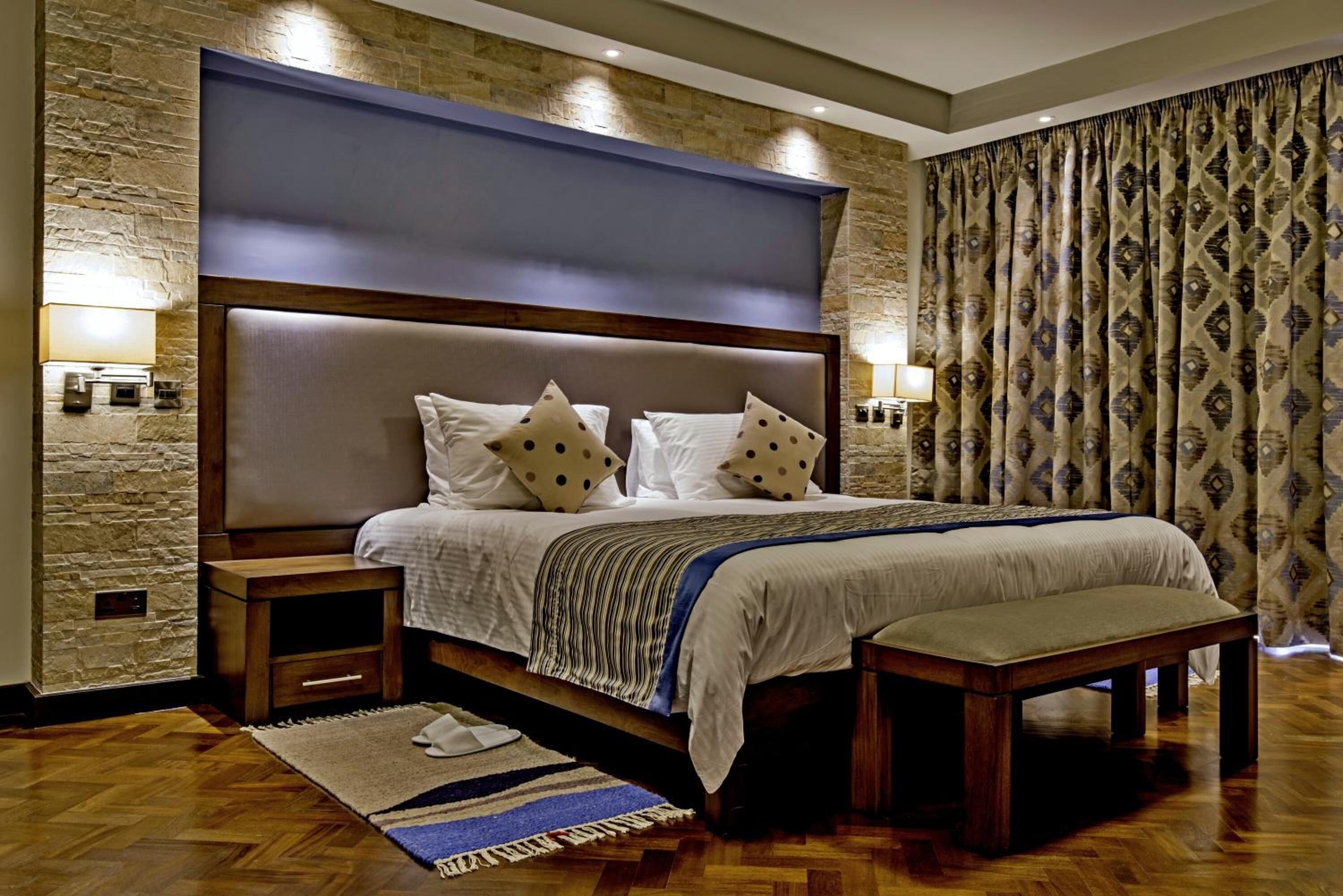 Un dormitorio con una cama grande, mesitas de noche de madera, un banco, una pared de piedra y cortinas estampadas.