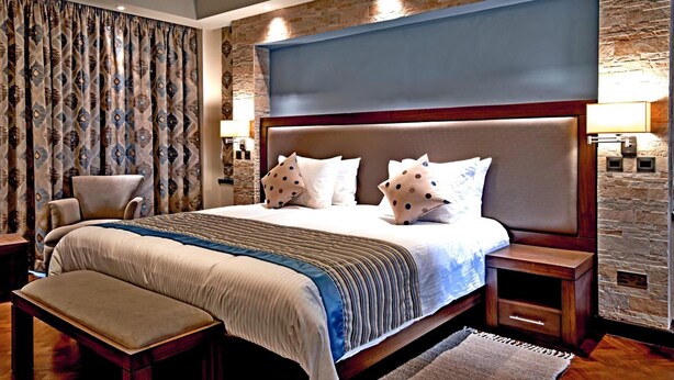 A modern bedroom with a large bed, a bench, a chair, and a stone accent wall. - Magna Hotel and Suites (Nairobi)