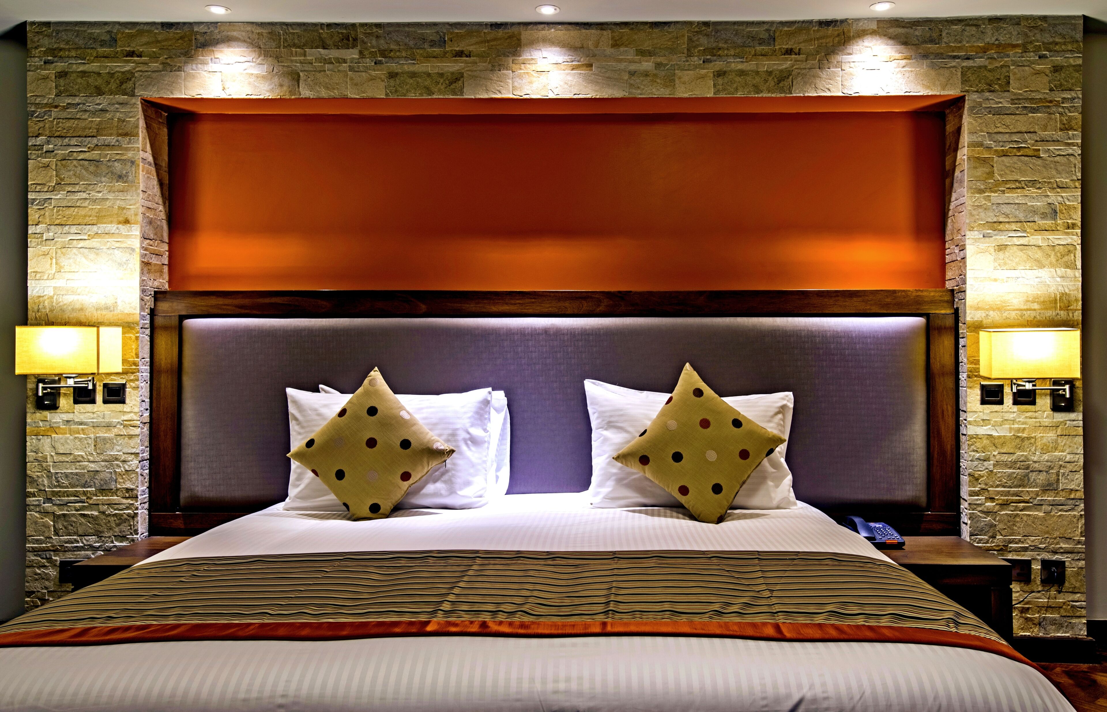 Quarto de hotel com parede de pedra, uma cama grande com roupa de cama branca e almofadas estampadas, e duas luminárias de cabeceira.