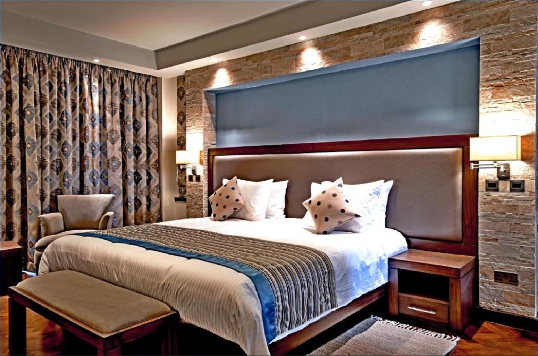 Quarto com cama grande, cabeceira, banqueta, cadeira e parede de pedra.