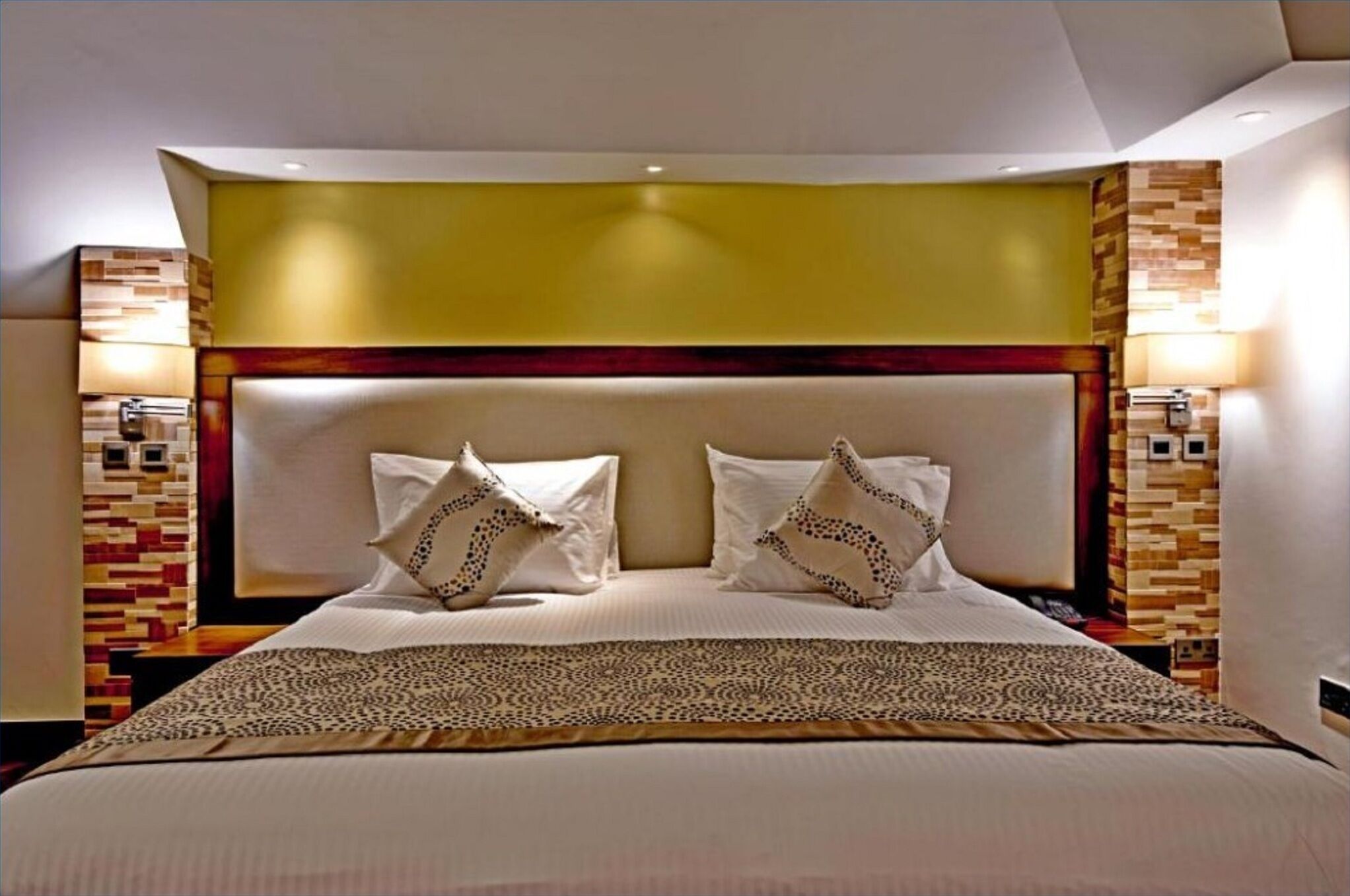 Quarto de hotel com cama grande, duas luminárias de cabeceira, cabeceira com padrão geométrico e manta decorativa.