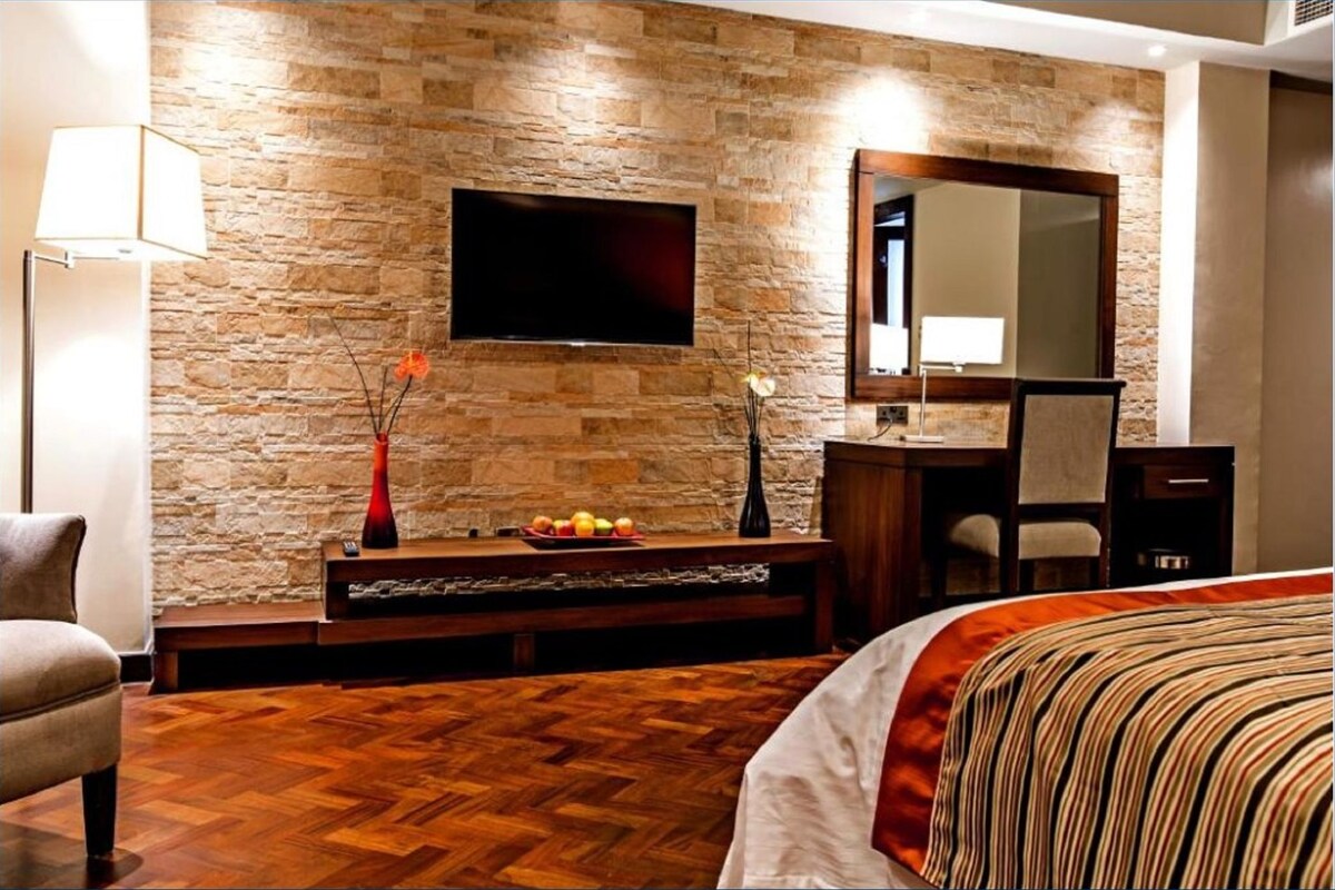 Habitación de hotel con pared de piedra, televisor de pantalla plana, banco de madera con frutero, cama con colcha a rayas y espejo sobre el banco.