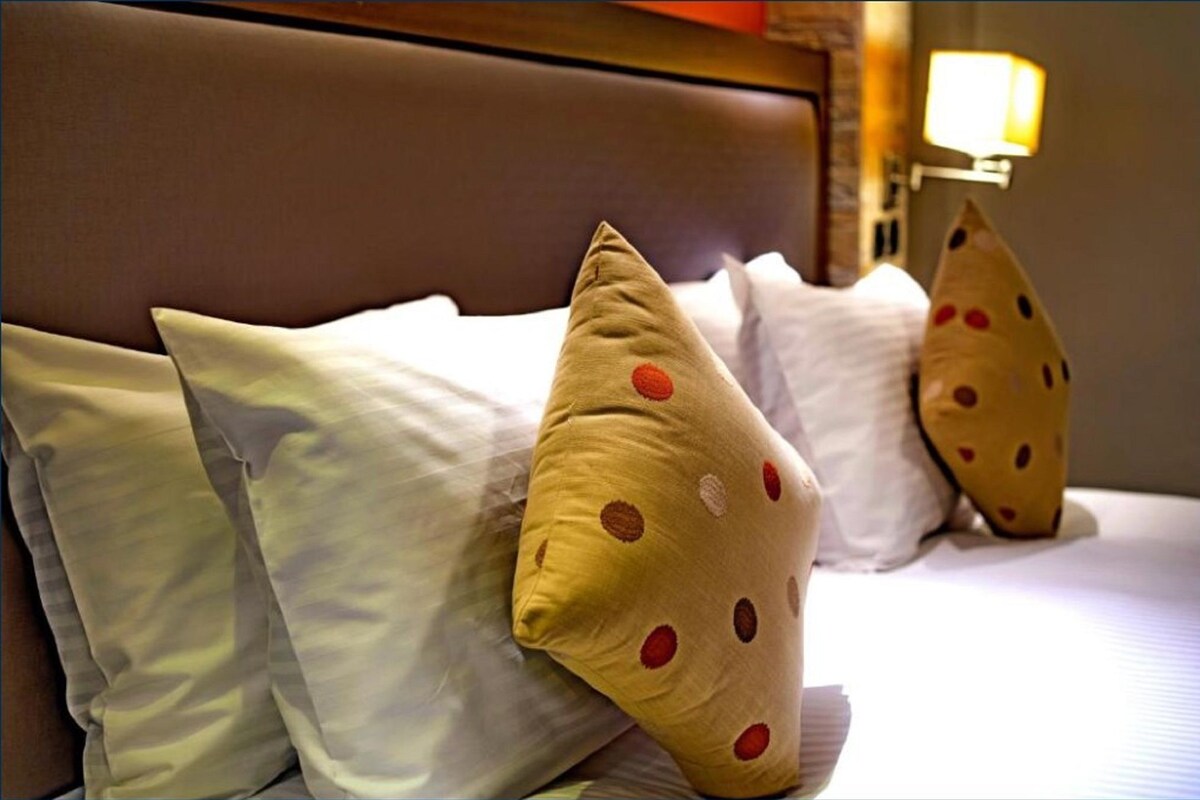 A bed with white and polka-dotted pillows, a brown headboard, and a wall-mounted lamp.