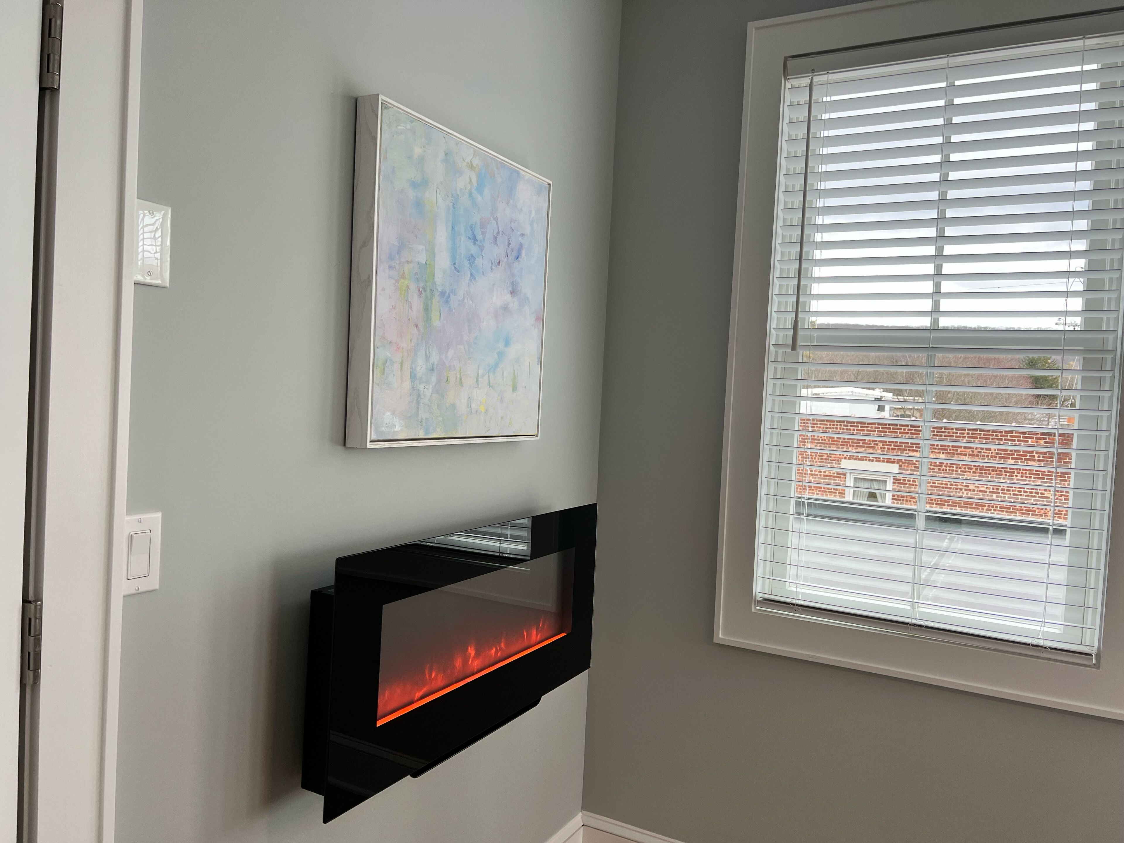 A modern living room with a fireplace, a window with blinds, and a piece of abstract art on the wall.