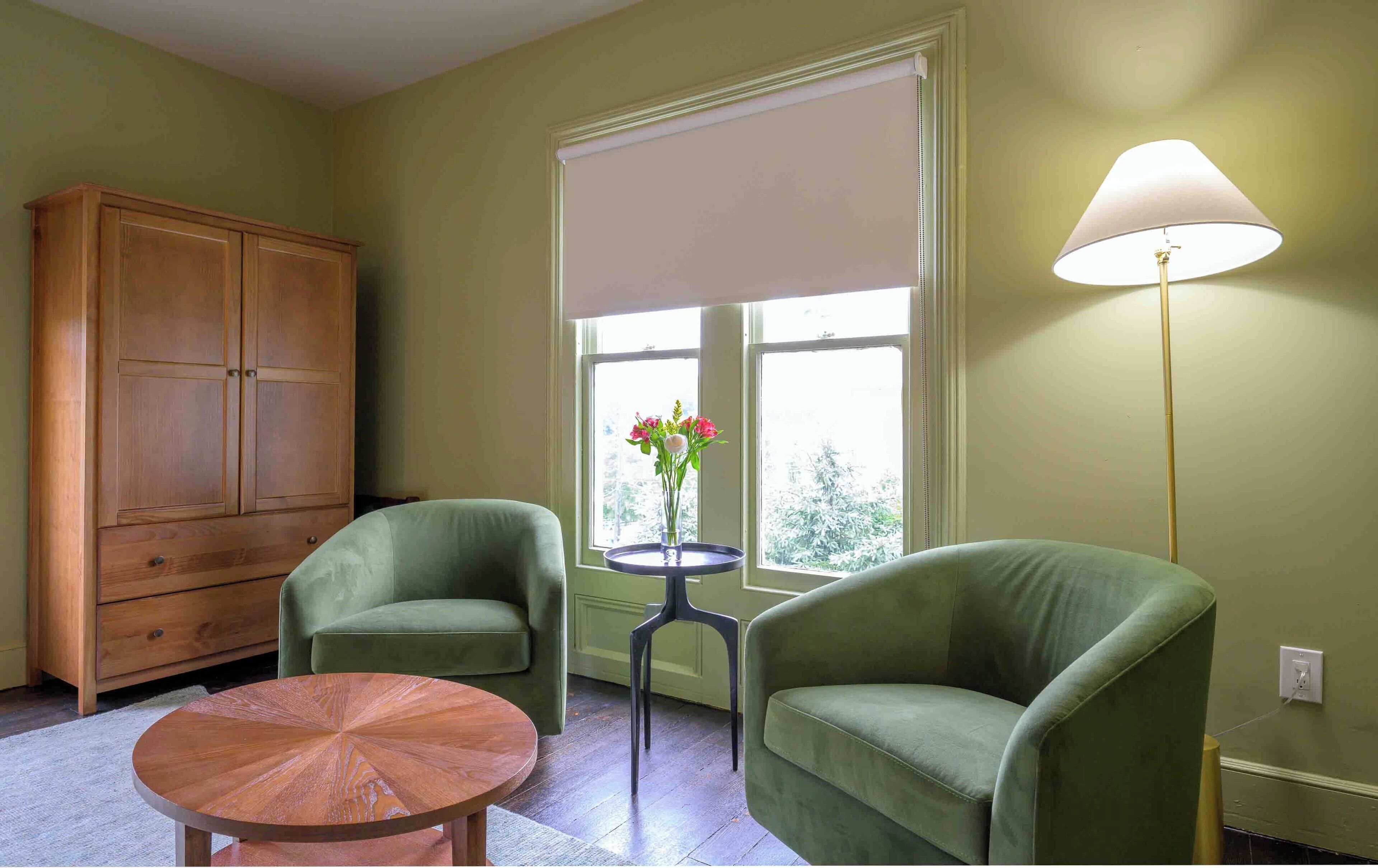 A room with two green armchairs, a wooden cabinet, a small round table, and a floor lamp.
