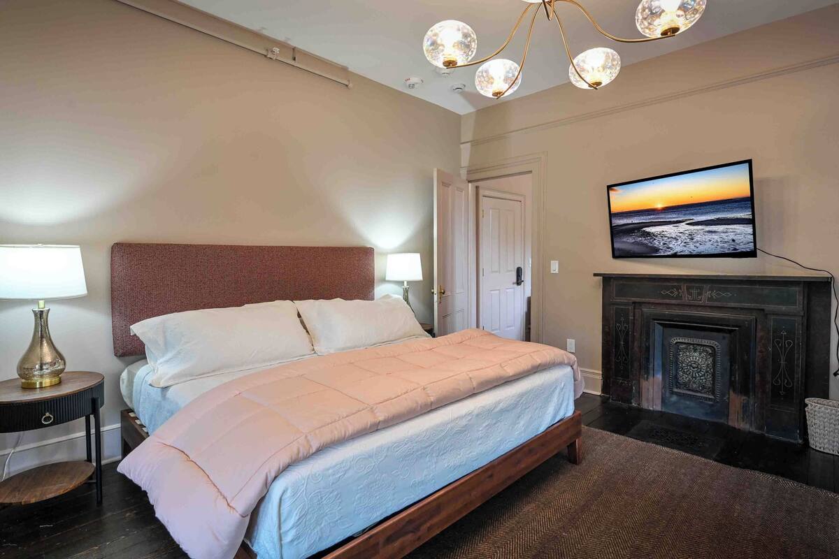 A bedroom with a bed, a television mounted on the wall, a fireplace, and a chandelier.