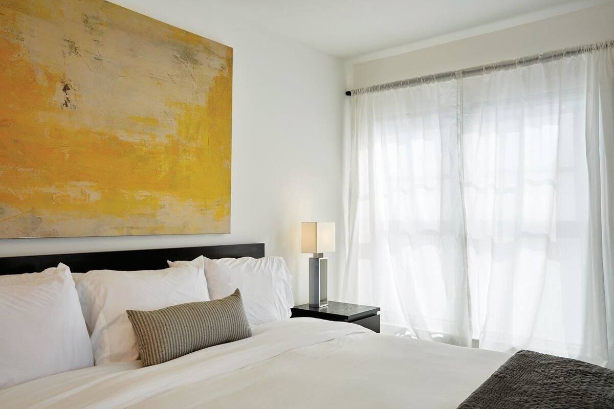 A hotel room with a large bed, white bedding, a nightstand with a lamp, and a large abstract painting on the wall.