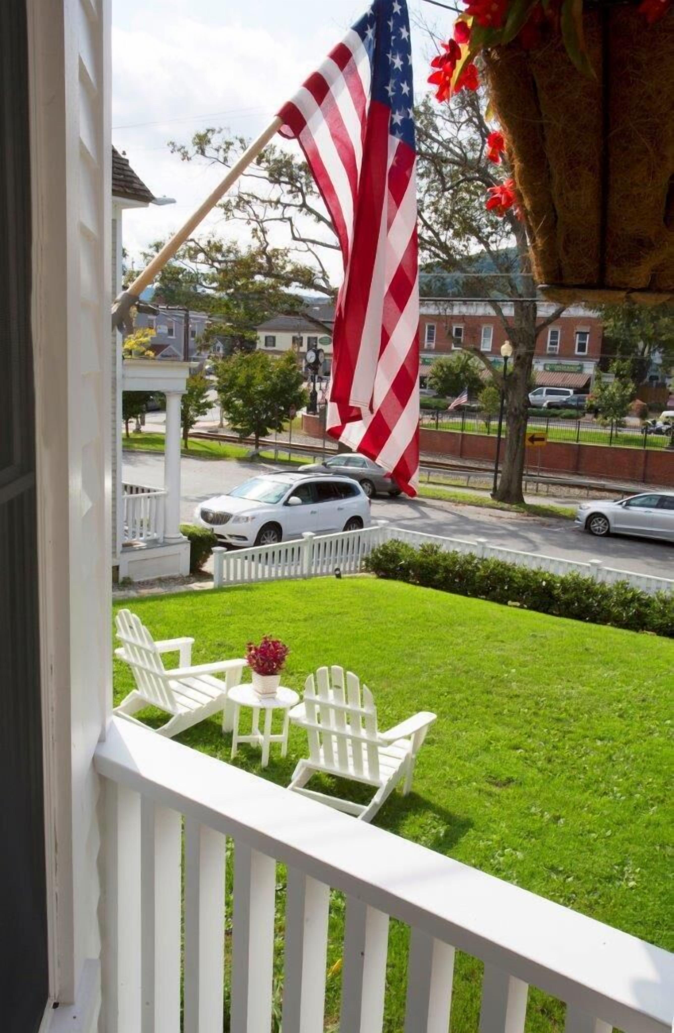 Eine Veranda mit weißen Stühlen und einem kleinen Tisch, eine amerikanische Flagge und der Blick auf eine Straße mit Autos und Häusern.