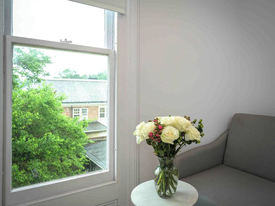 Een raam met uitzicht op een gebouw en bomen, een vaas met witte bloemen op een klein tafeltje, en een grijze bank.
