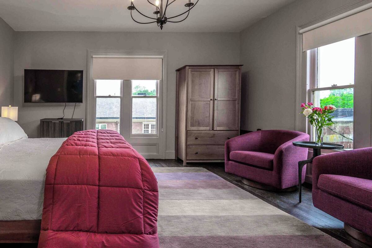 A bedroom with a bed, a wardrobe, a TV, a chair, and a table with flowers.