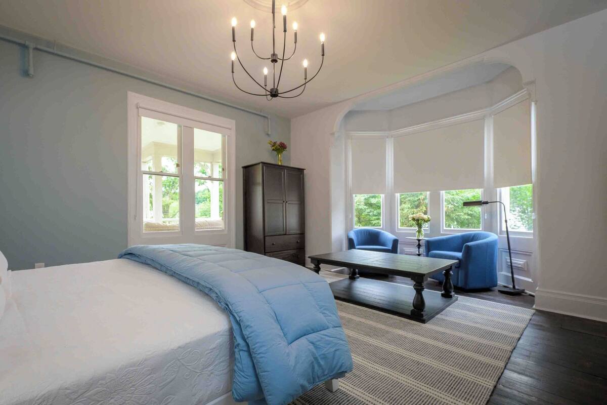 A bedroom with a large bed, a chandelier, a dark wooden cabinet, and a seating area with two chairs and a table.