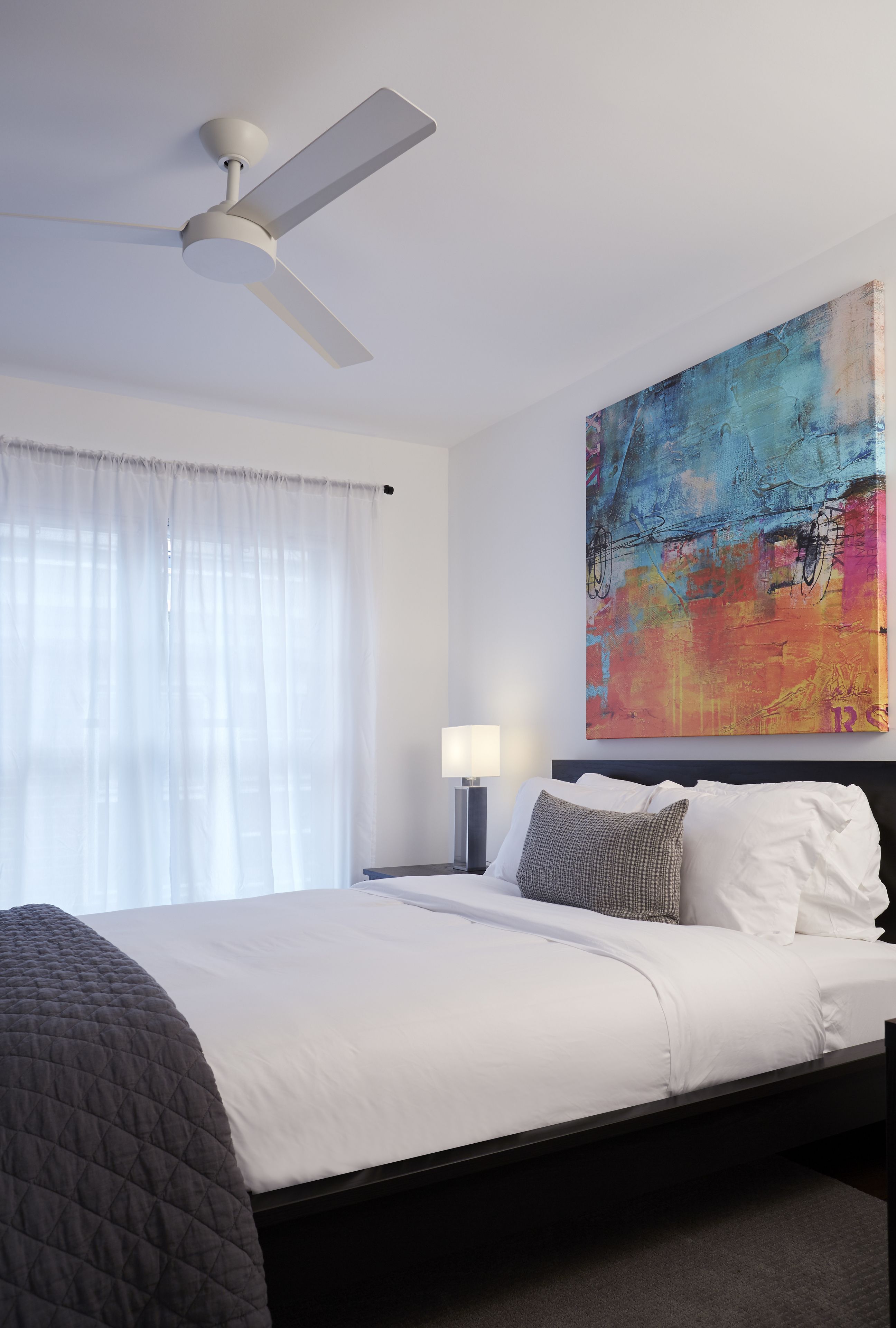 A hotel room with a bed, a ceiling fan, a painting, a nightstand, and a lamp.