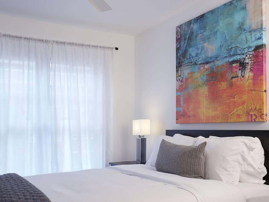 A hotel room with a bed, a ceiling fan, a painting, a nightstand, and a lamp.