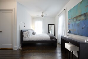 A bedroom with a bed, a desk, a chair, a ceiling fan, a large abstract painting, and a window with curtains.