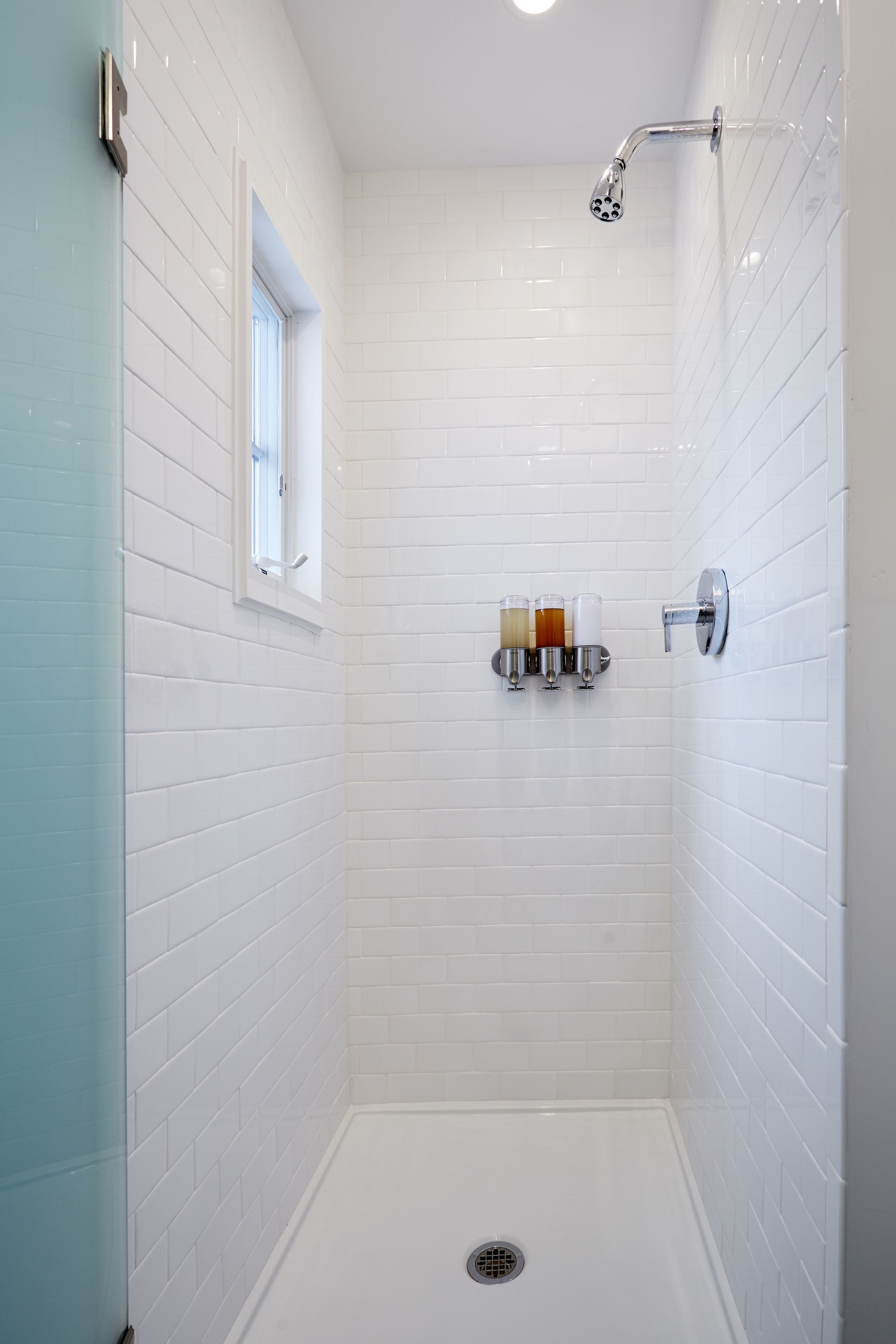 Pequeño cuarto de ducha con azulejos blancos, ventana, cabezal de ducha y estante con artículos de aseo.