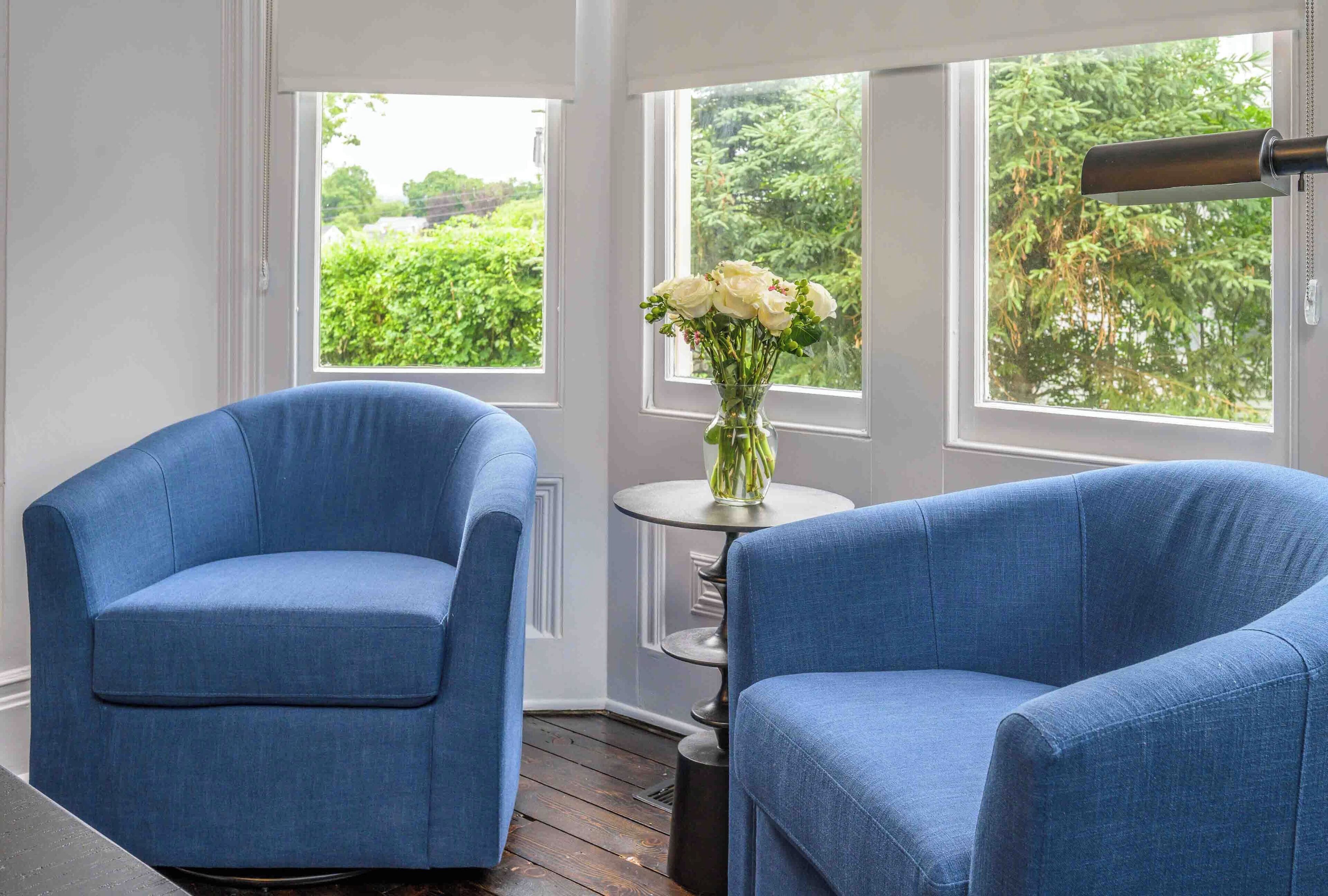A room with two blue armchairs, a small round table with a vase of white flowers, and large windows with a view of greenery.