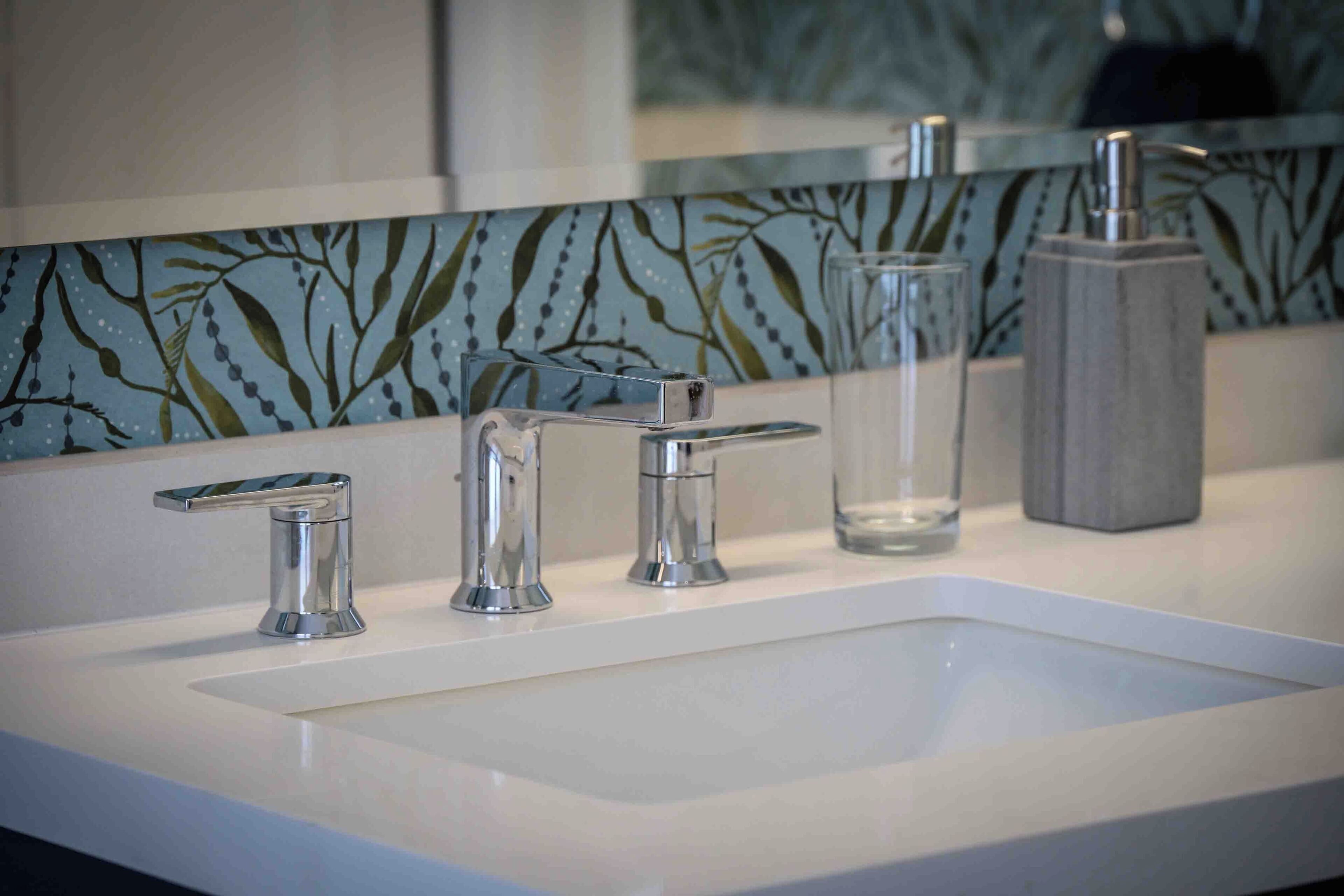 A modern bathroom with a white sink, two chrome faucets, and a glass container.