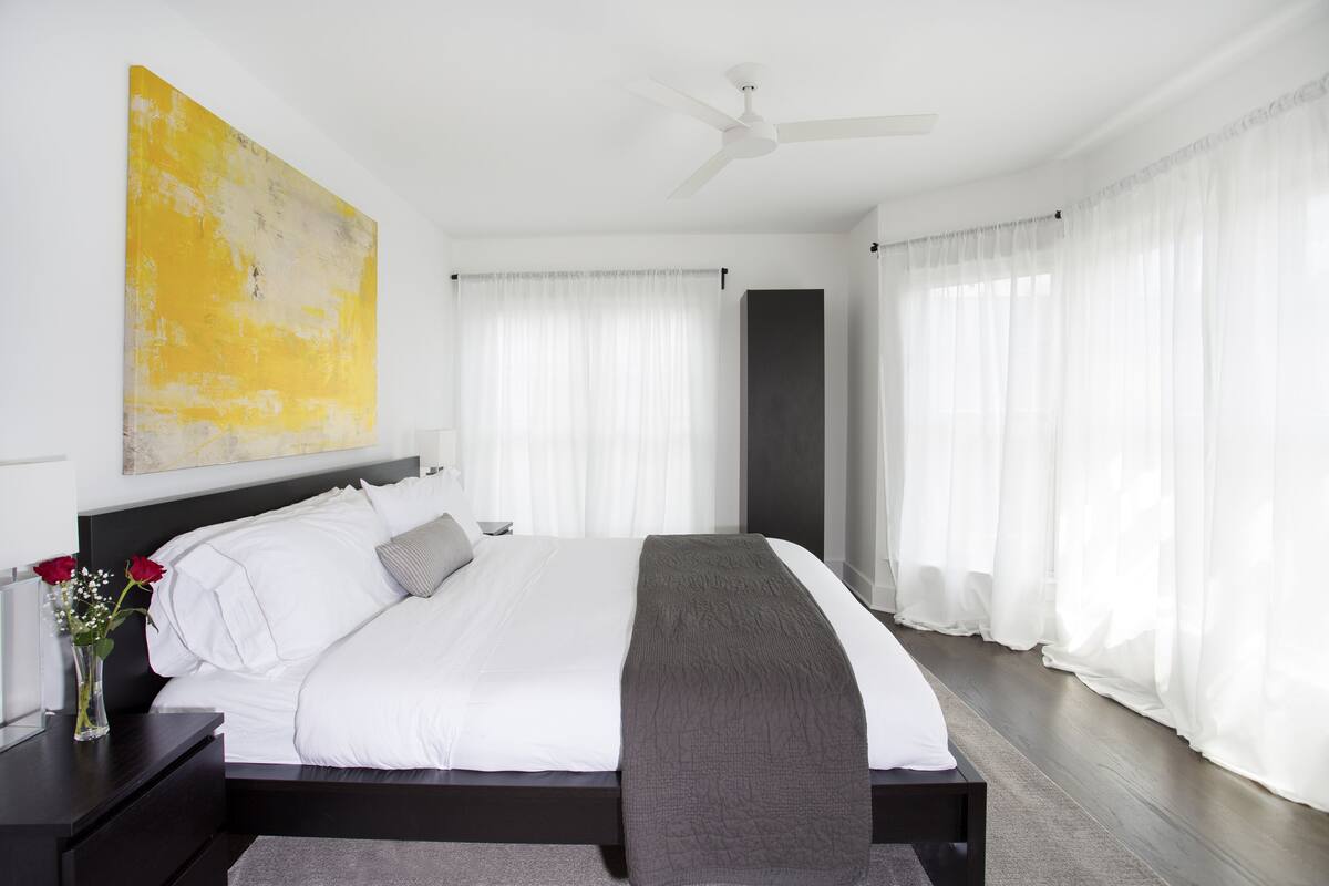 A modern bedroom with a large bed, white bedding, a gray throw blanket, and a yellow abstract painting on the wall.