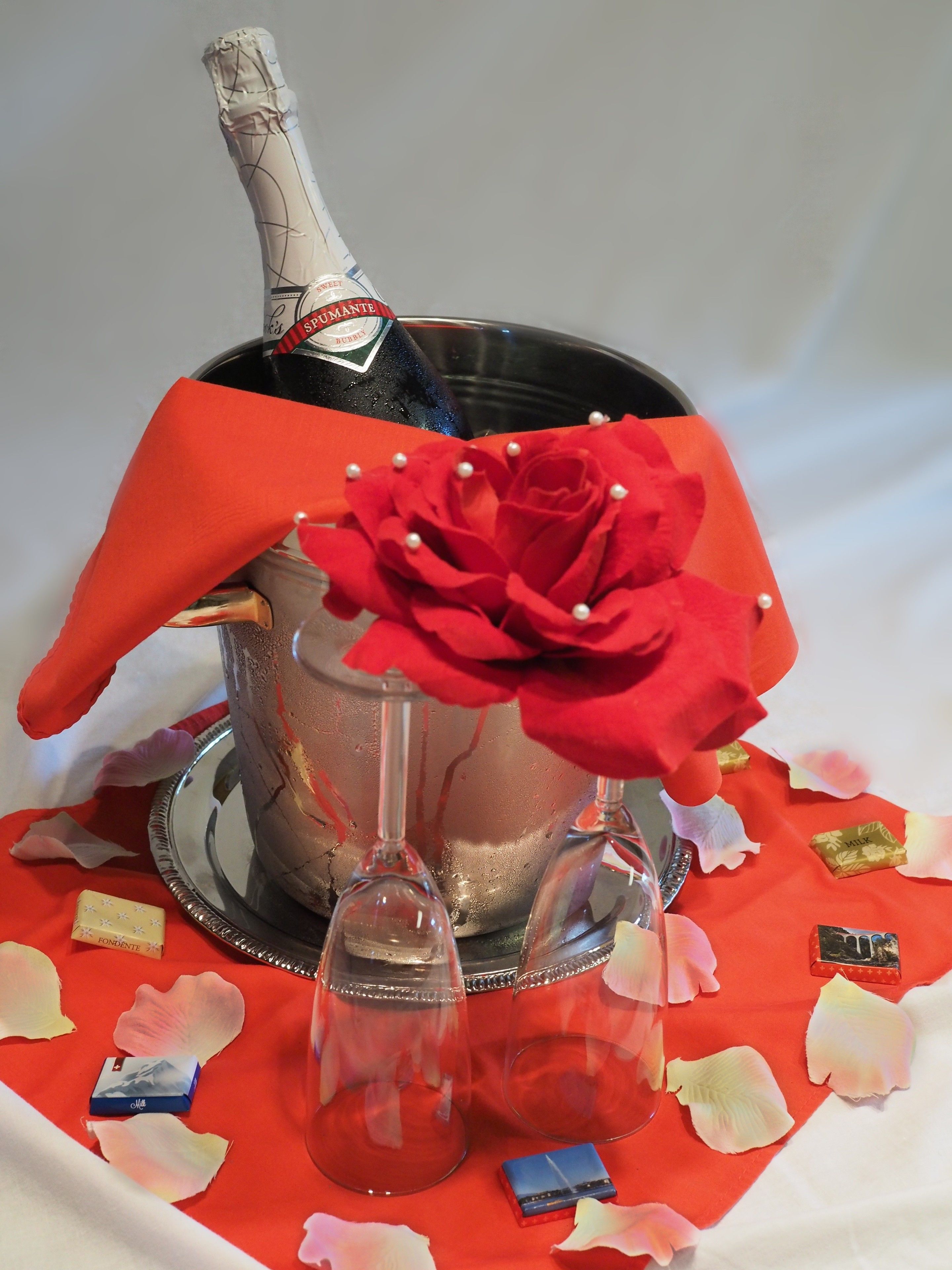 A champagne bottle in a silver bucket with a red cloth and a red rose decoration.
