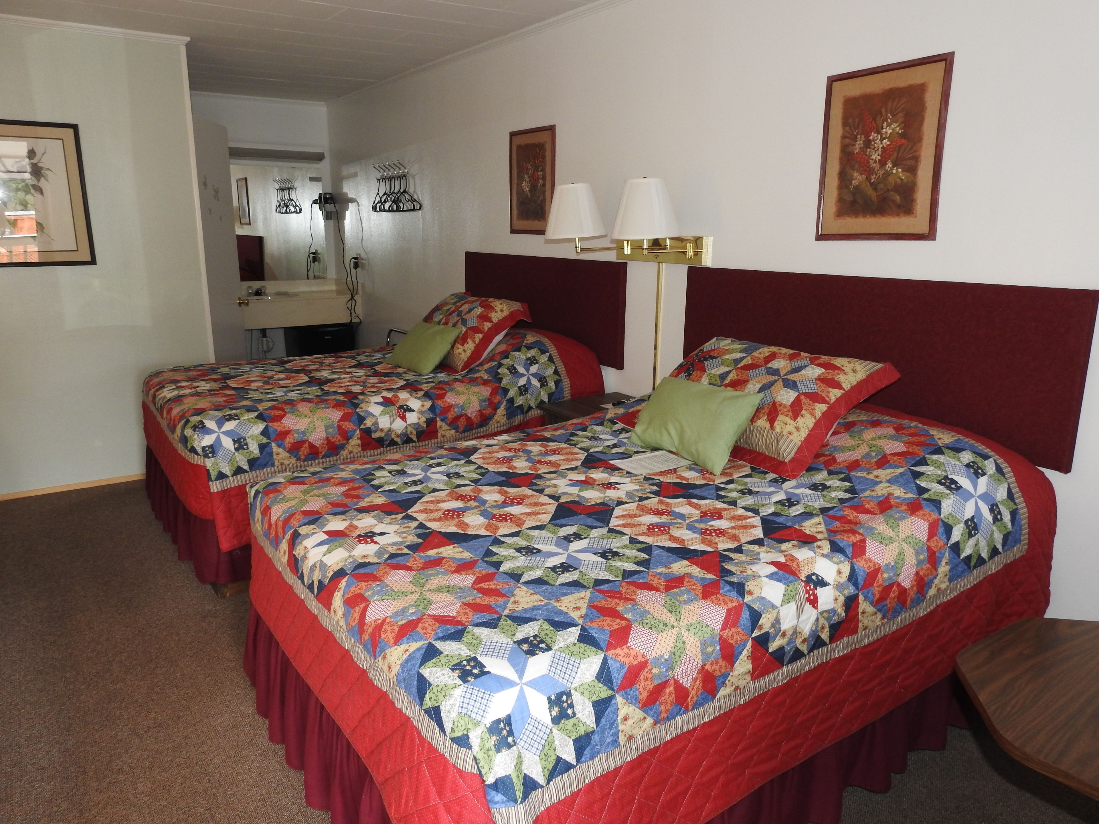 A hotel room with two beds, each with a colorful quilt, a bedside table, a lamp, and framed artwork on the walls.