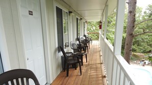 A covered porch with wooden flooring, white railings, and several black plastic chairs.