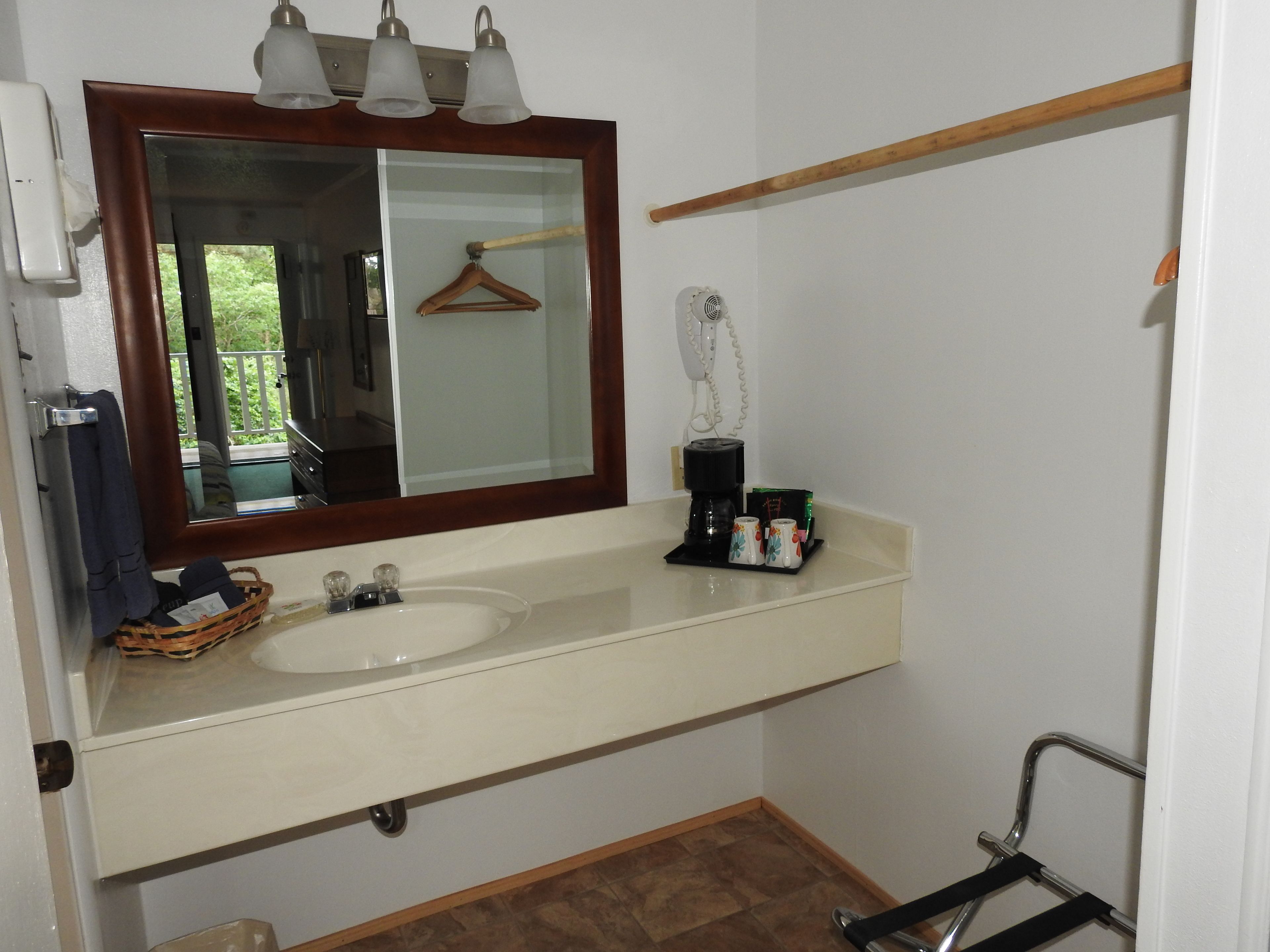 A bathroom with a double sink, a mirror with lights, a towel rack, a hanging coat, a coffee maker, and a shower chair.