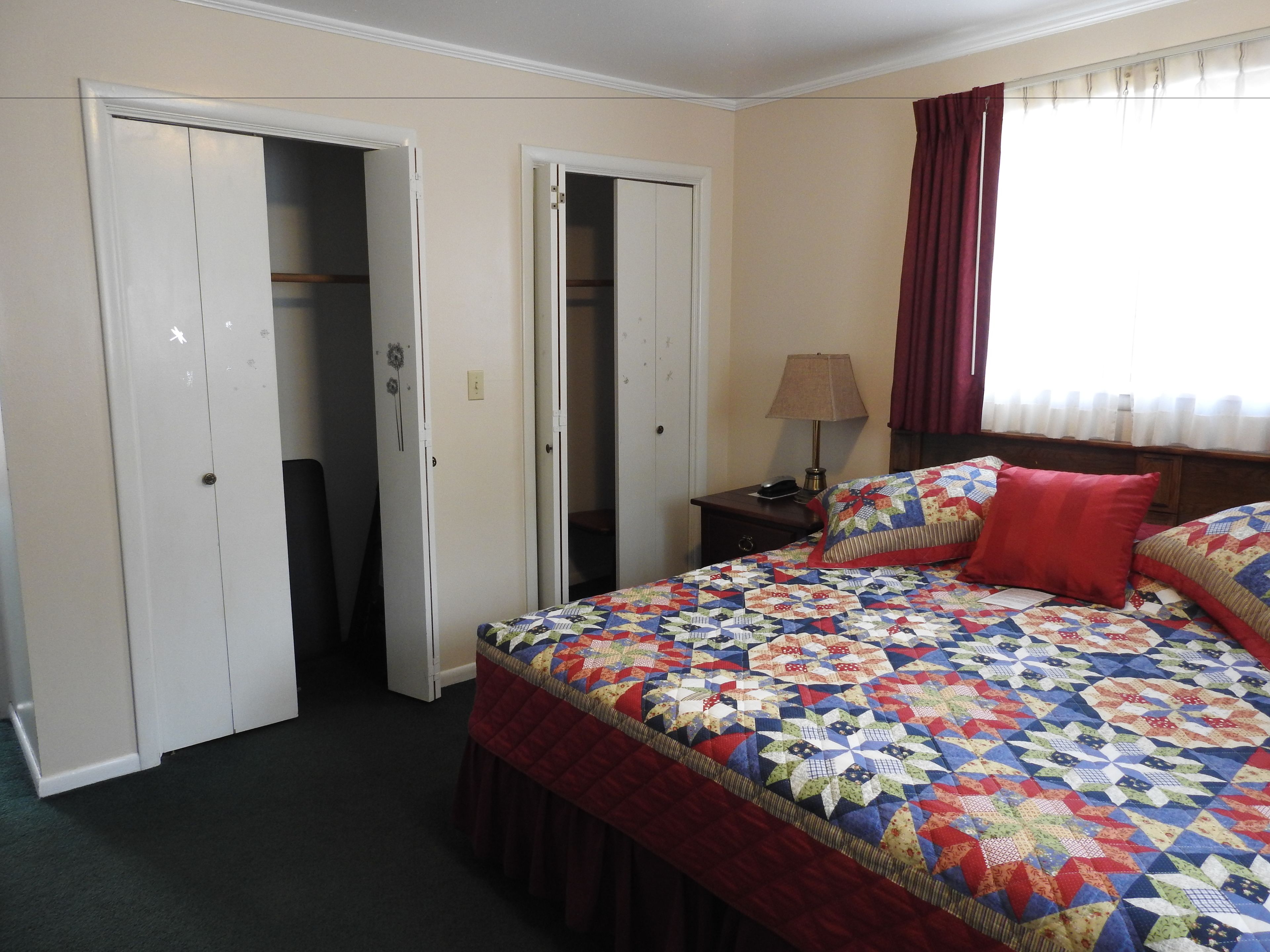 Quarto com cama grande, dois armários abertos, mesa de cabeceira com abajur e janela com cortinas.