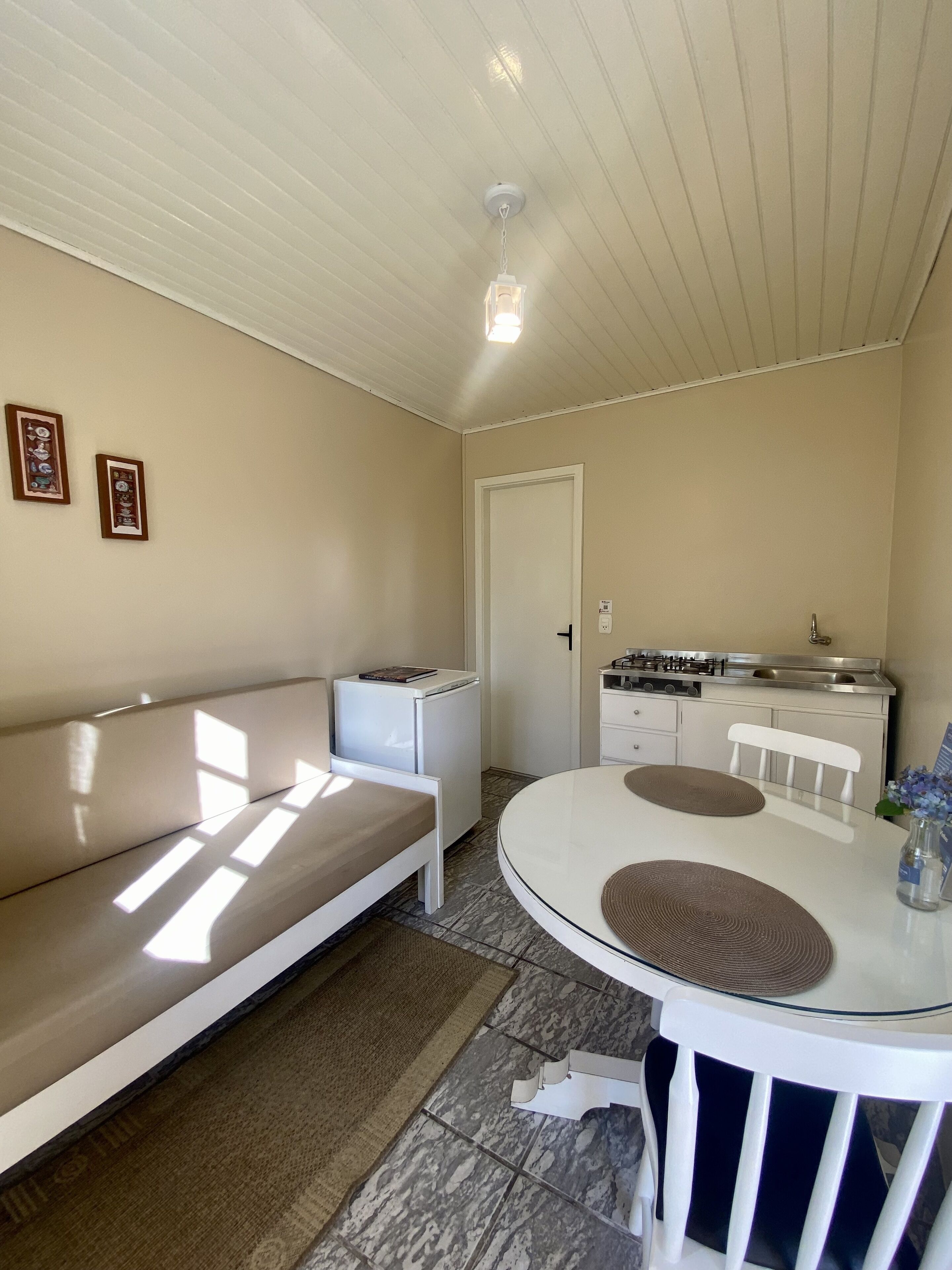 A compact living space with a white sofa, a small kitchenette, a dining table with chairs, and a ceiling light fixture.
