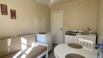 A compact living space with a white sofa, a small kitchenette, a dining table with chairs, and a ceiling light fixture.