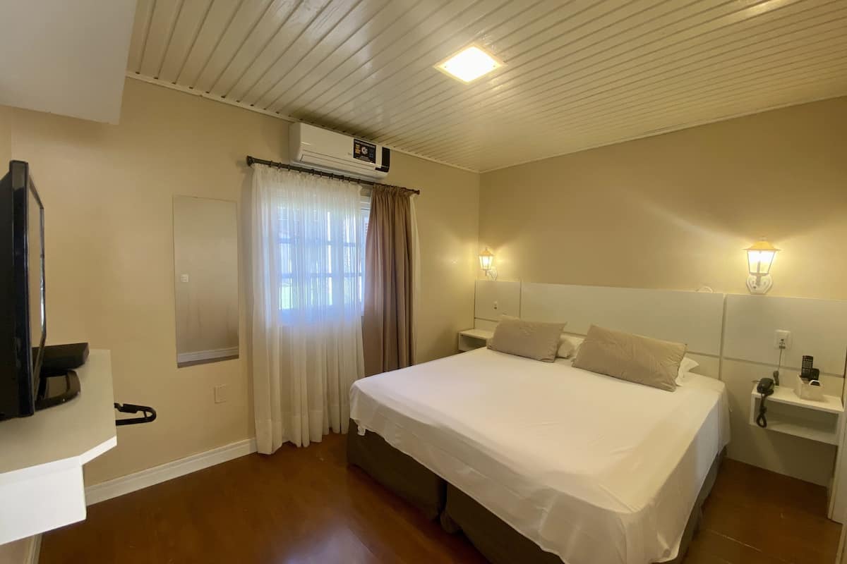 A hotel room with a bed, a TV, an air conditioning unit, a window with curtains, and two wall-mounted light fixtures.