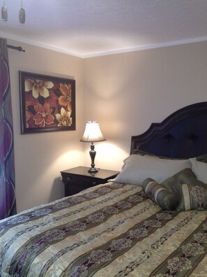 Ein ordentlich bezogenes Bett mit einem gemusterten Überwurf, einer Nachttischlampe und einem floralen Bild an der Wand.