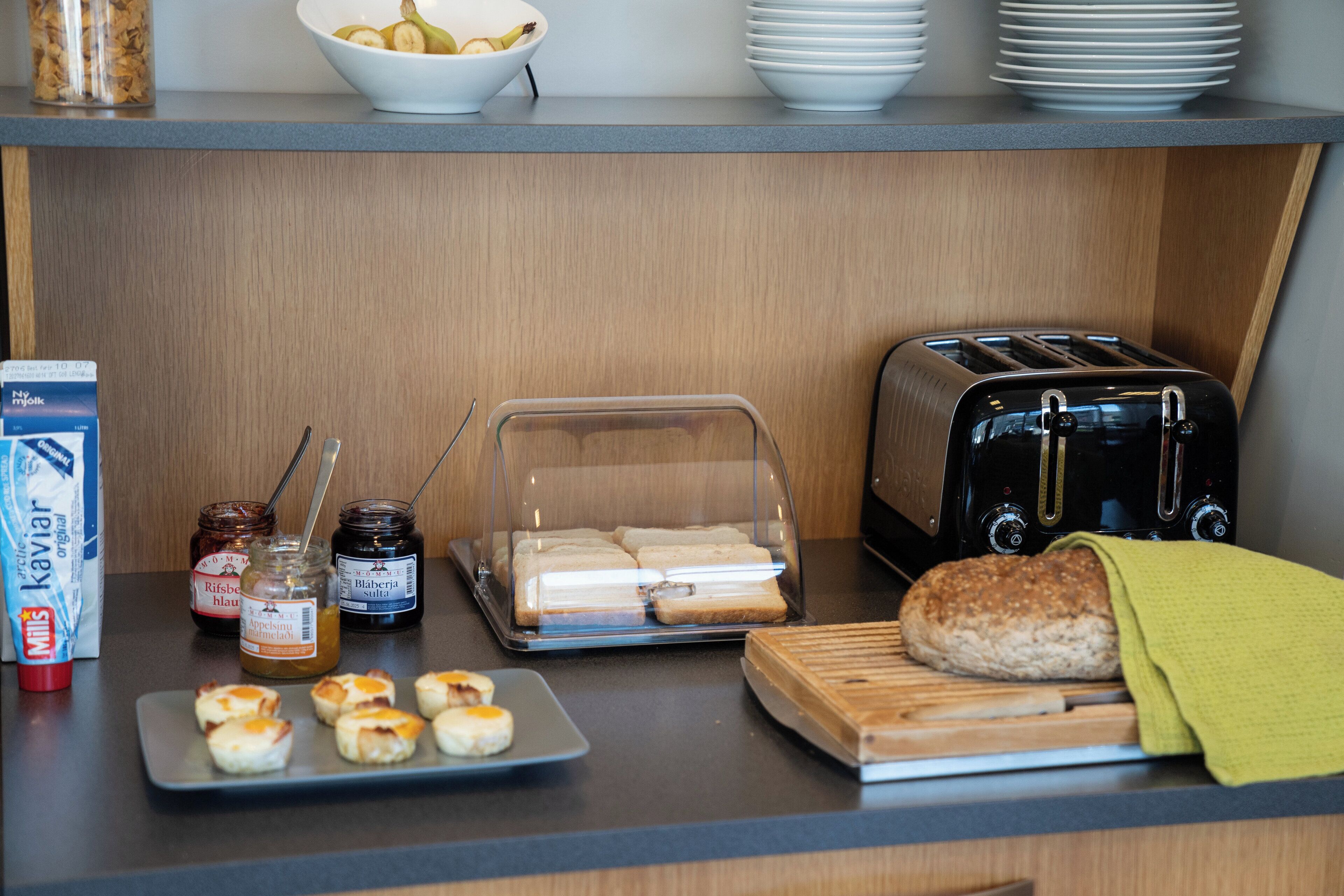 A breakfast setup with a toaster, bread, a cutting board, and a tray of pastries.