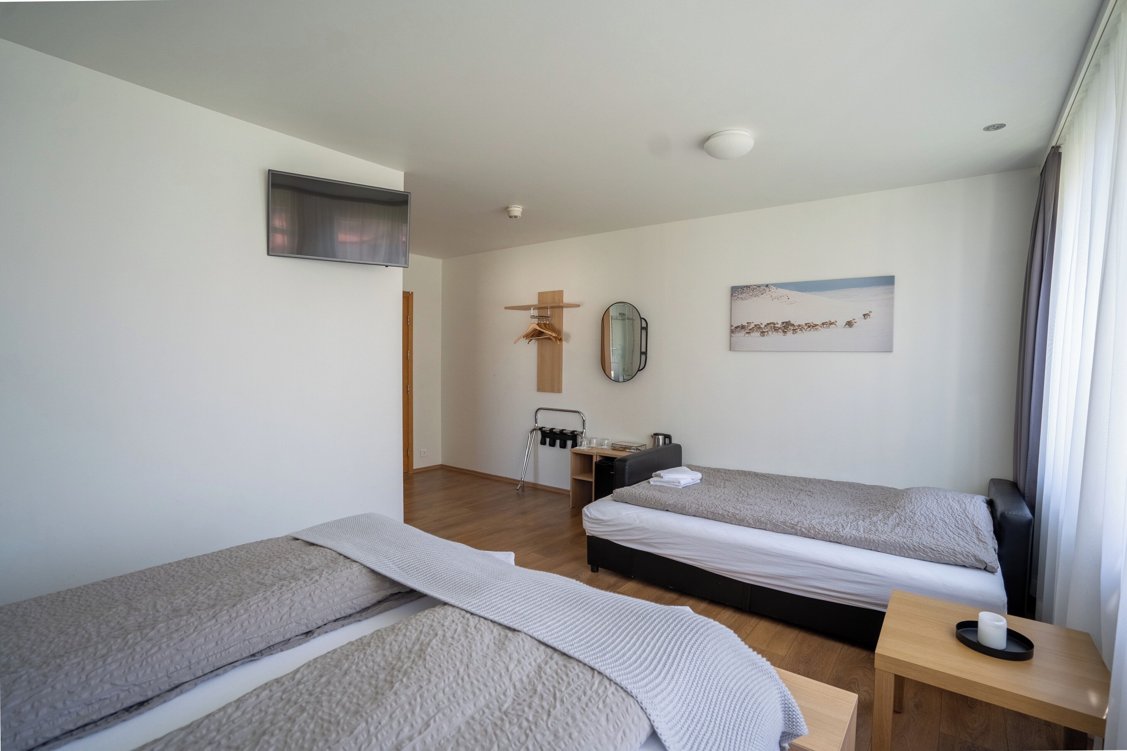 A modern hotel room with two single beds, a wooden side table, a small wooden stool, a wall-mounted TV, a mirror, and a painting.