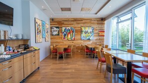 Een moderne eetruimte met houten wanden en een groot raam. Er staan verschillende tafels en stoelen, en er hangt kunst aan de muren.